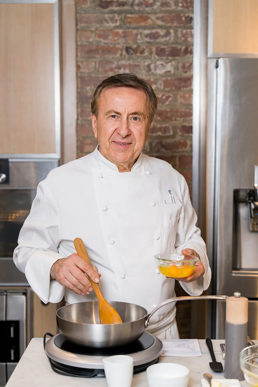 A senior male chef in a white uniform stands in a kitchen, holding a bowl of raw eggs in one hand and a wooden spoon in the other over a stove.