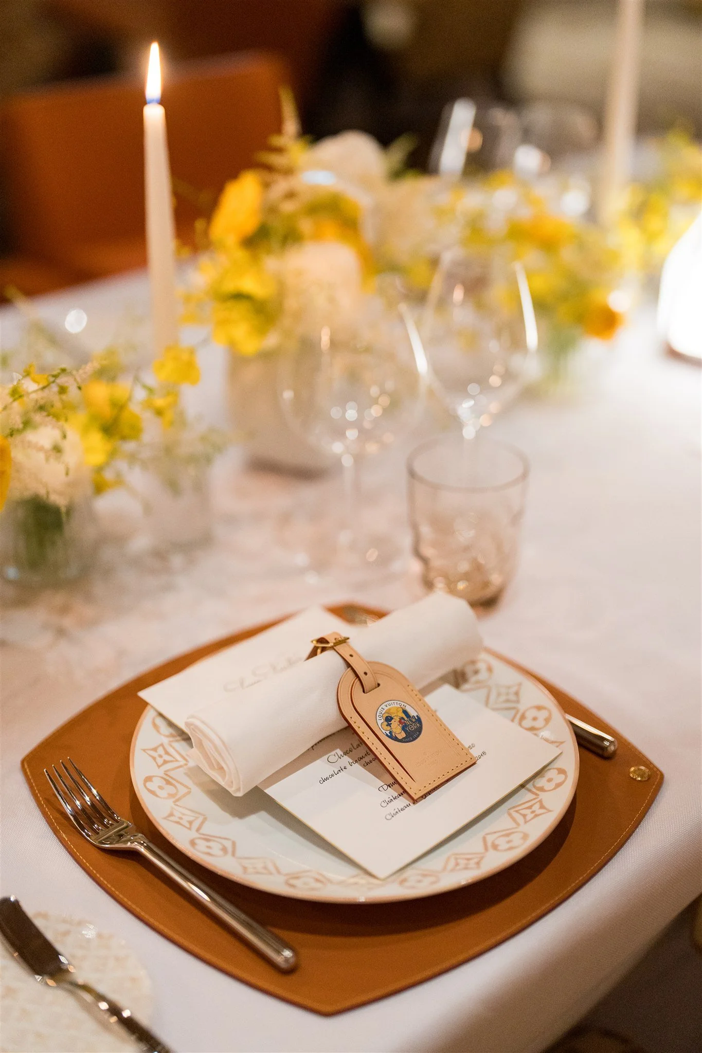 Elegant dinner table setting with a white plate, a menu, a rolled napkin, a place card, silverware, wine glasses, and a gold-colored placemat, with floral centerpieces and candles in the background.