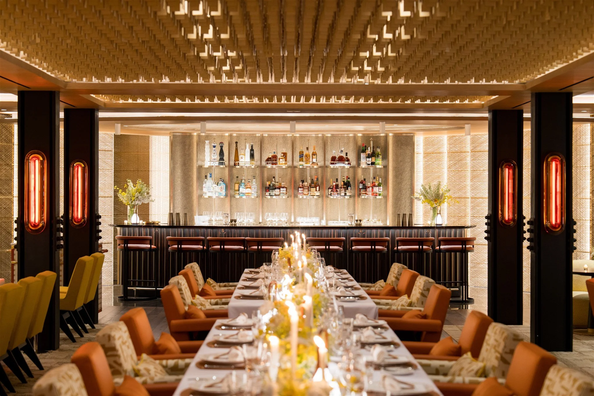 Elegant restaurant with a long dining table set with candles and flowers, a bar in the background with alcohol bottles and glassware, decorative wall lighting, and modern seating arrangements.