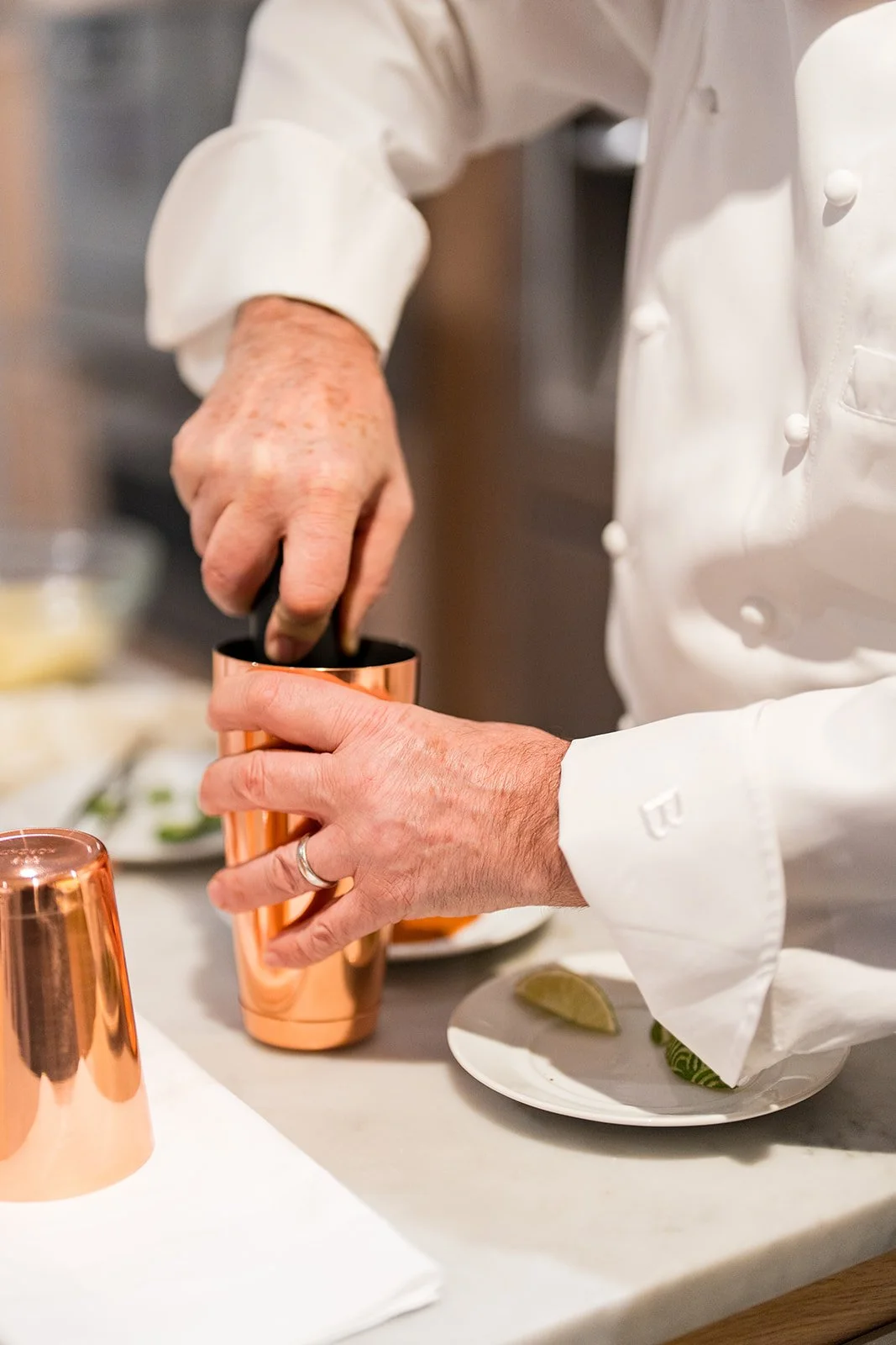 Chef in a white coat preparing a cocktail, holding a copper plating shaker and pouring an ingredient into a glass.
