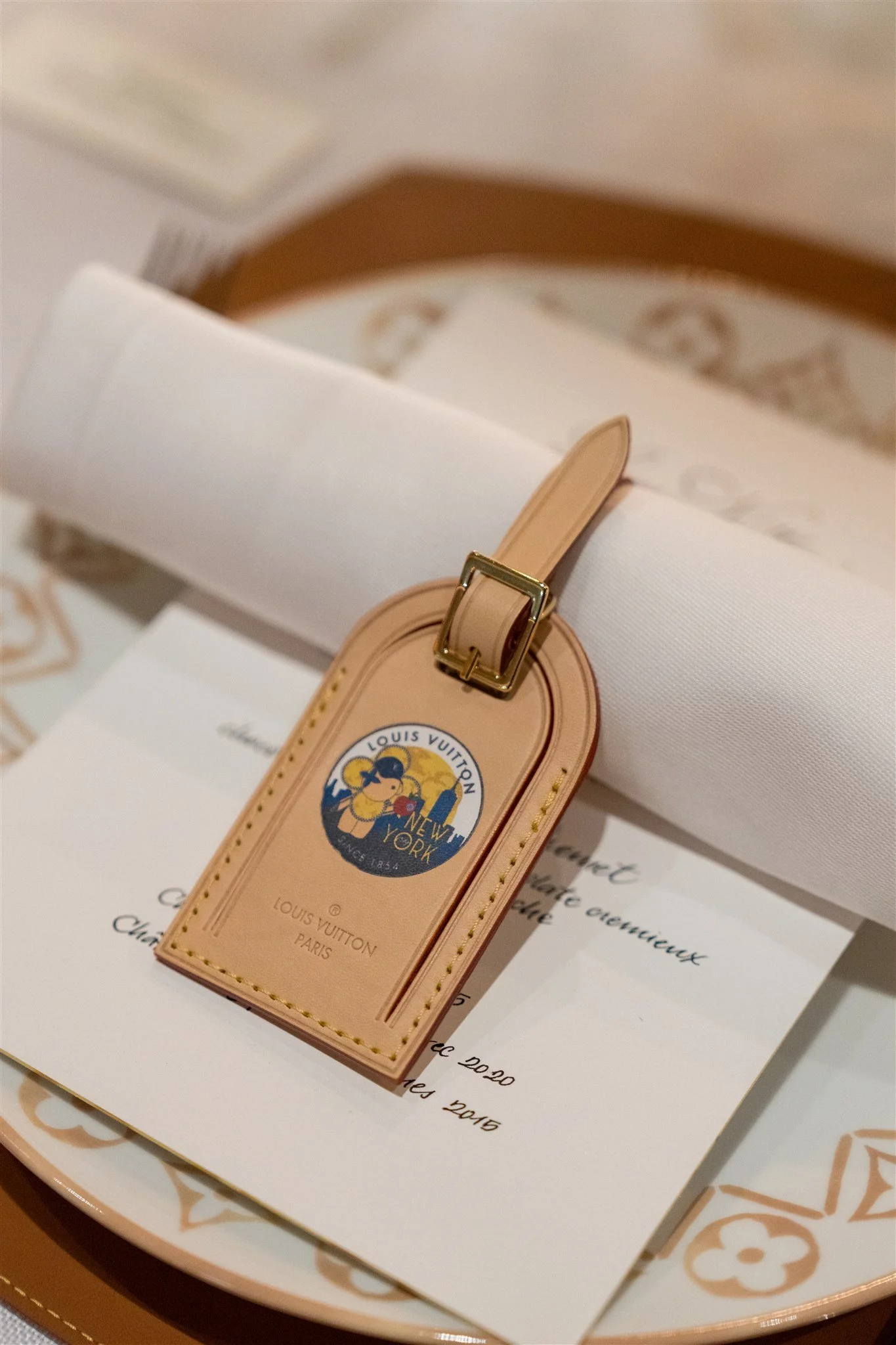 A Louis Vuitton luggage tag with a Mickey Mouse design, placed on top of a white paper and a Louis Vuitton branded item in the background.