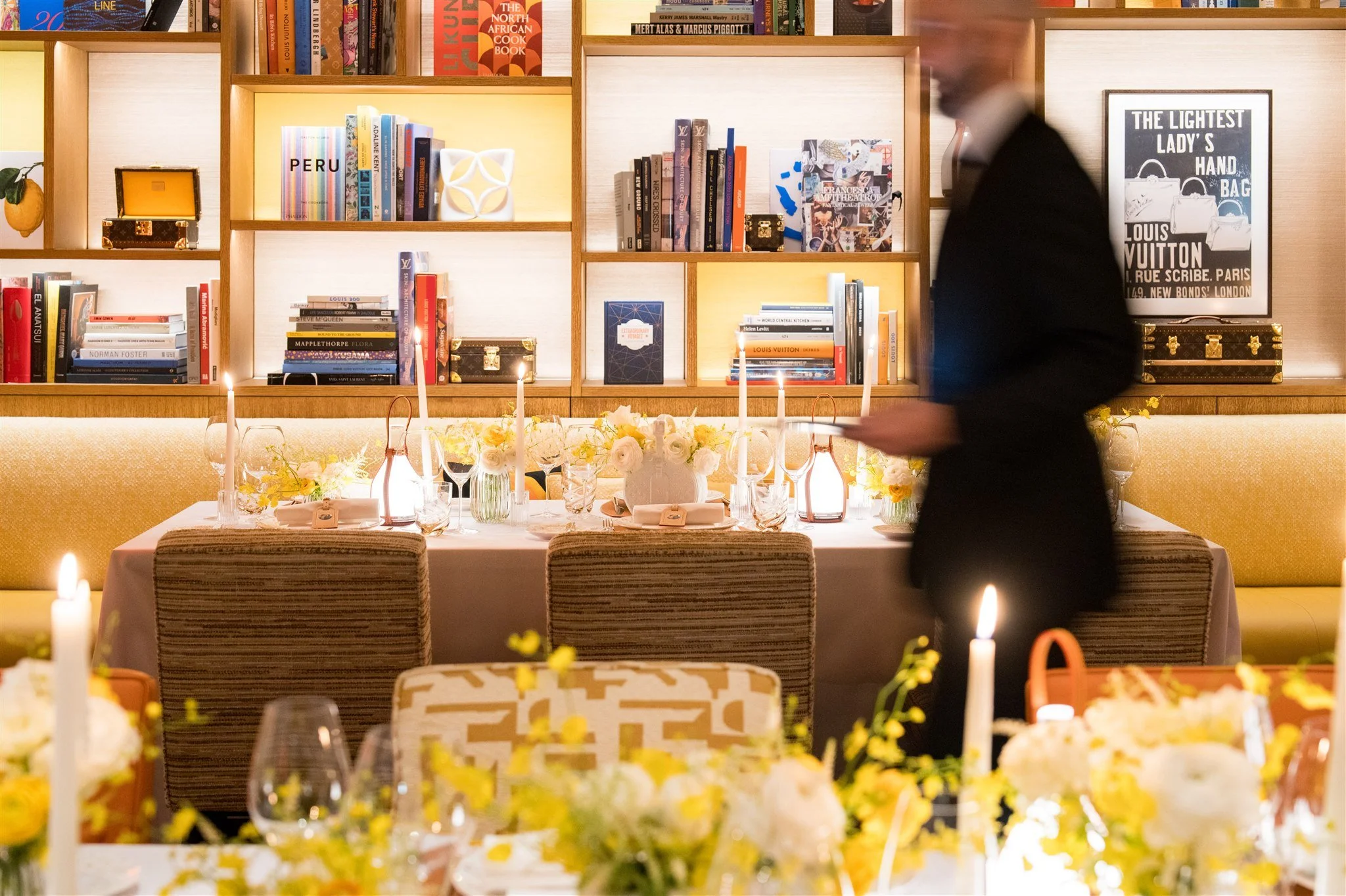 A dining room with a long table set for an event, decorated with candles and floral arrangements, with a person in a dark suit walking past. The background features a bookcase filled with books, decorative objects, and framed posters.