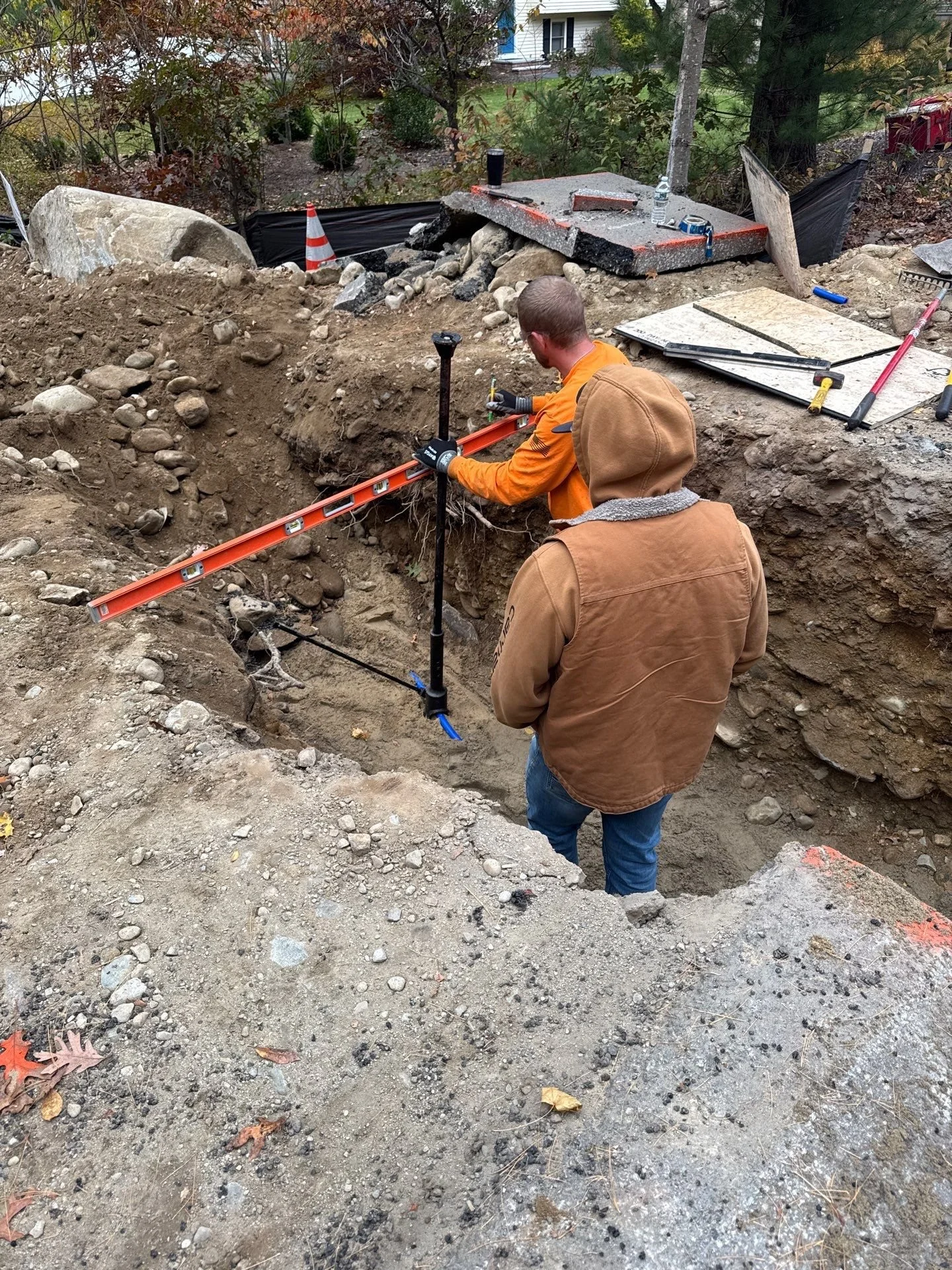 Two construction workers in a dugout hole at a construction site, measuring and leveling the ground with tools. One worker is using a level, and the other is observing.