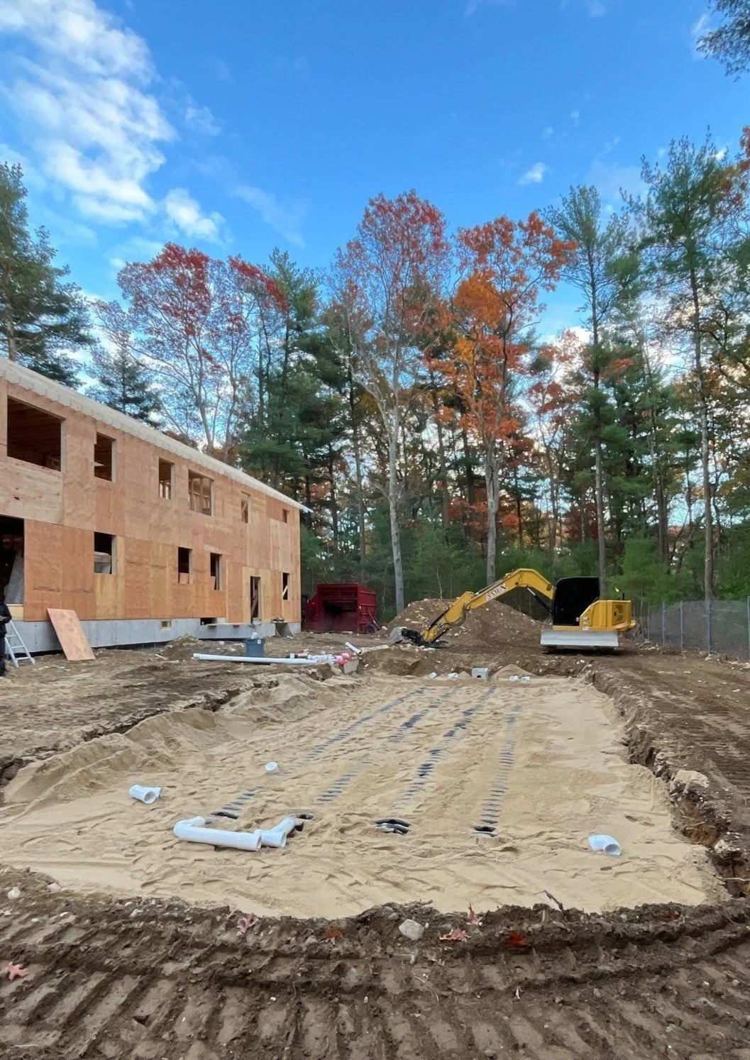 Construction site with an unfinished building, yellow excavator, and dirt ground, surrounded by trees with some fall foliage, under a partly cloudy sky.