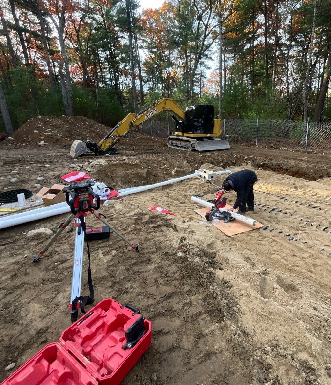 Construction site with a small excavator, a worker handling pipes, and equipment for laying underground pipes near a wooded area.
