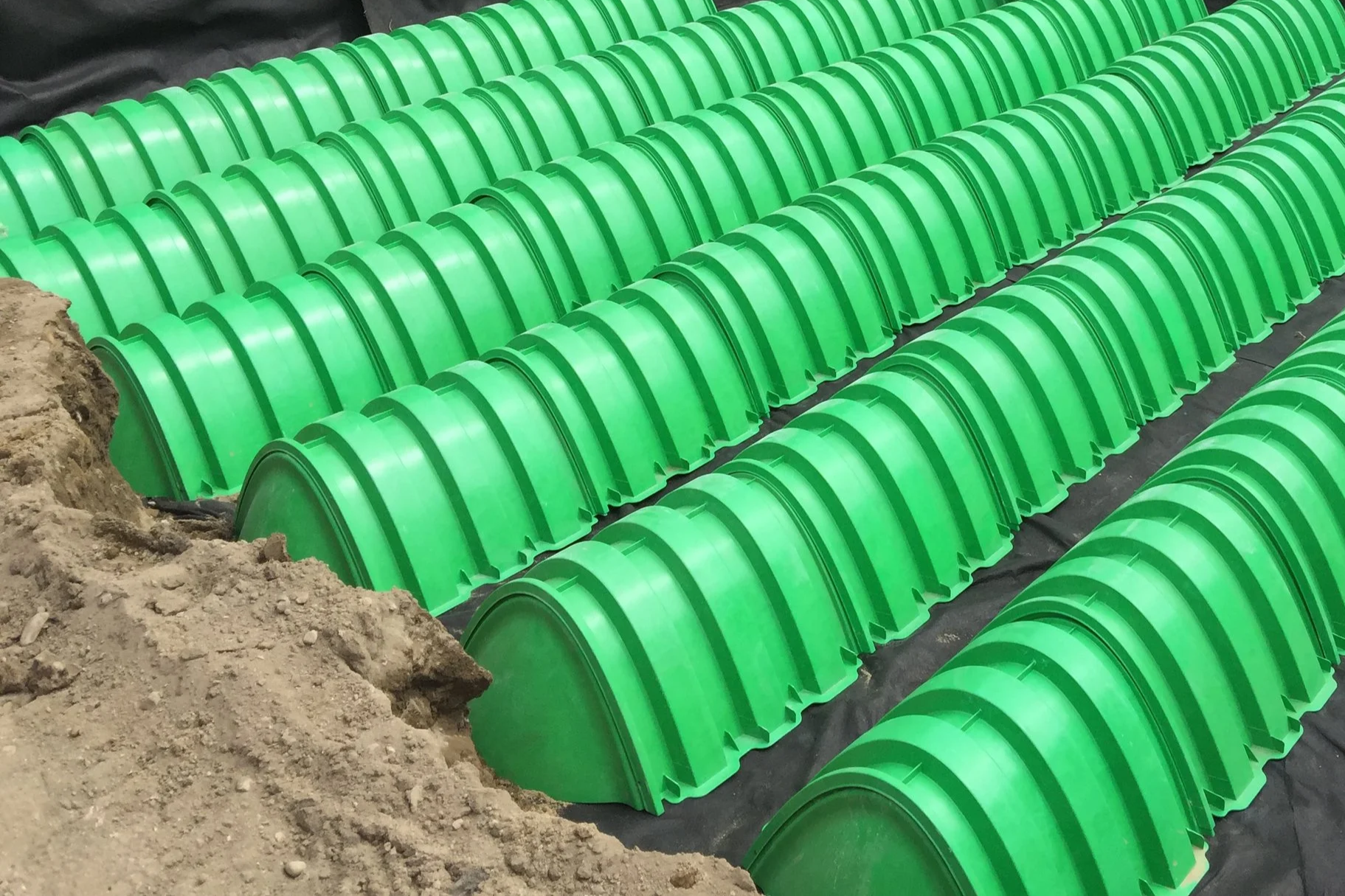 Rows of green plastic drainage pipes arranged in a construction site with dirt in the foreground.