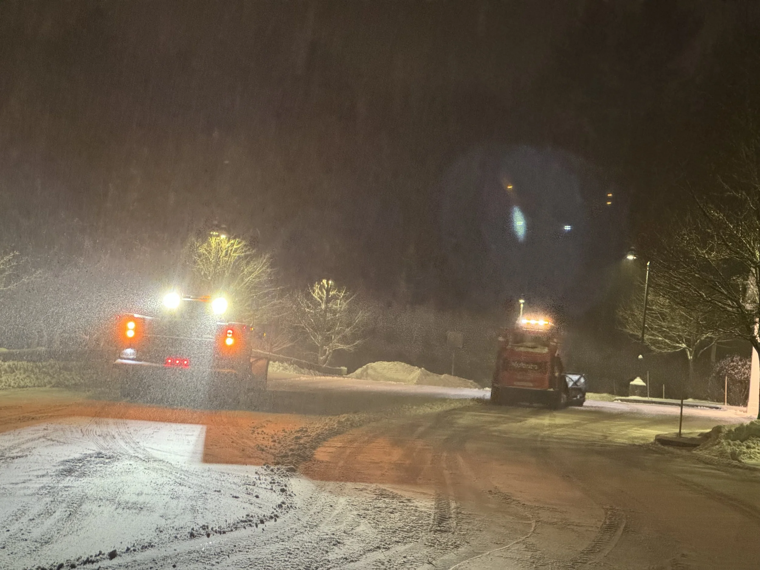 Nighttime scene of a construction site with a large yellow front loader and a white work truck on a wet parking lot, illuminated by bright artificial lights. Snow is falling, and bare trees are visible in the background.