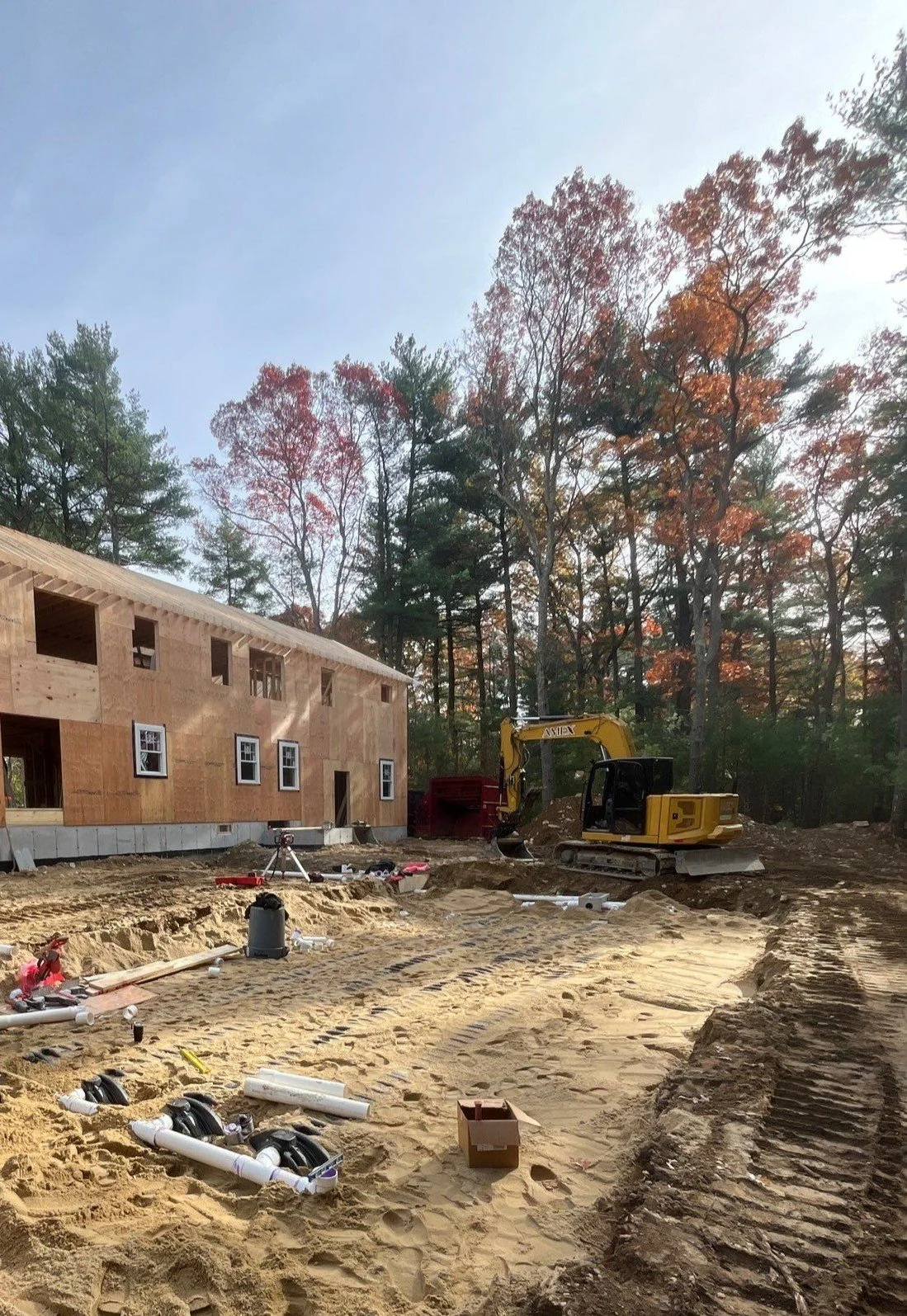 Construction site with a two-story wooden building under construction, a small yellow excavator, and scattered pipes and tools on sandy ground. Tall trees with autumn leaves are in the background.