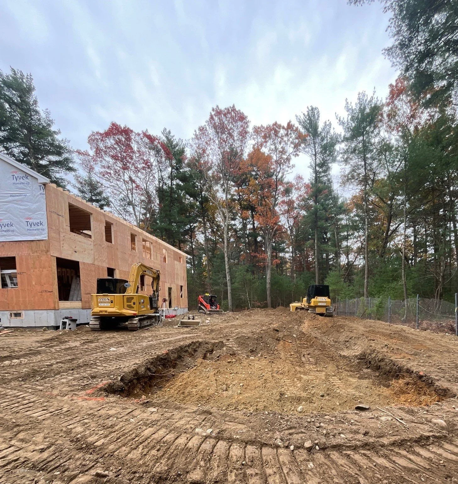 Construction site with a new multi-story building under construction, construction vehicles on dirt ground, and trees in the background.