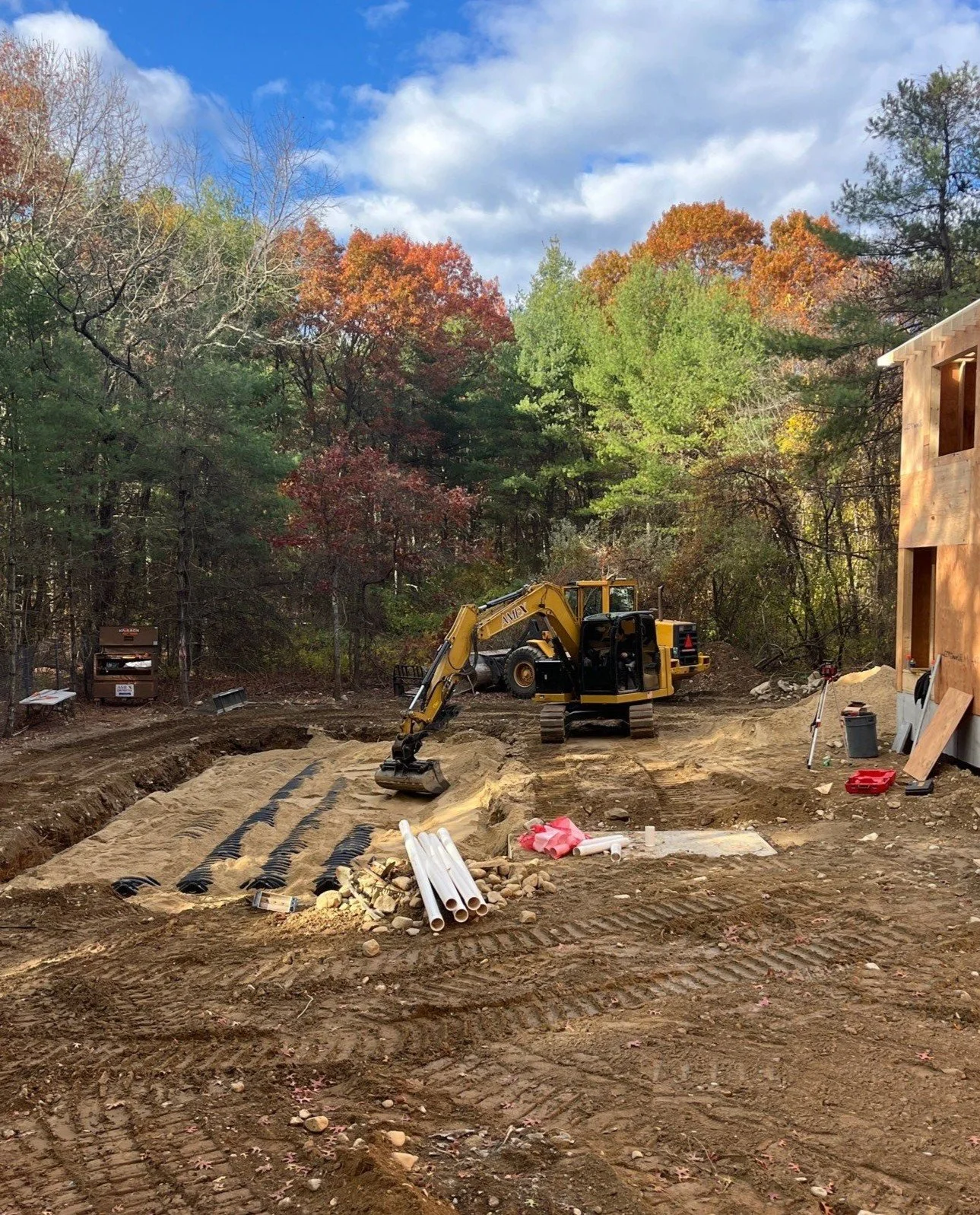 Construction site with a yellow excavator digging in the dirt, stacks of white pipes, and a partially built wooden structure surrounded by colorful trees under a blue sky with clouds.