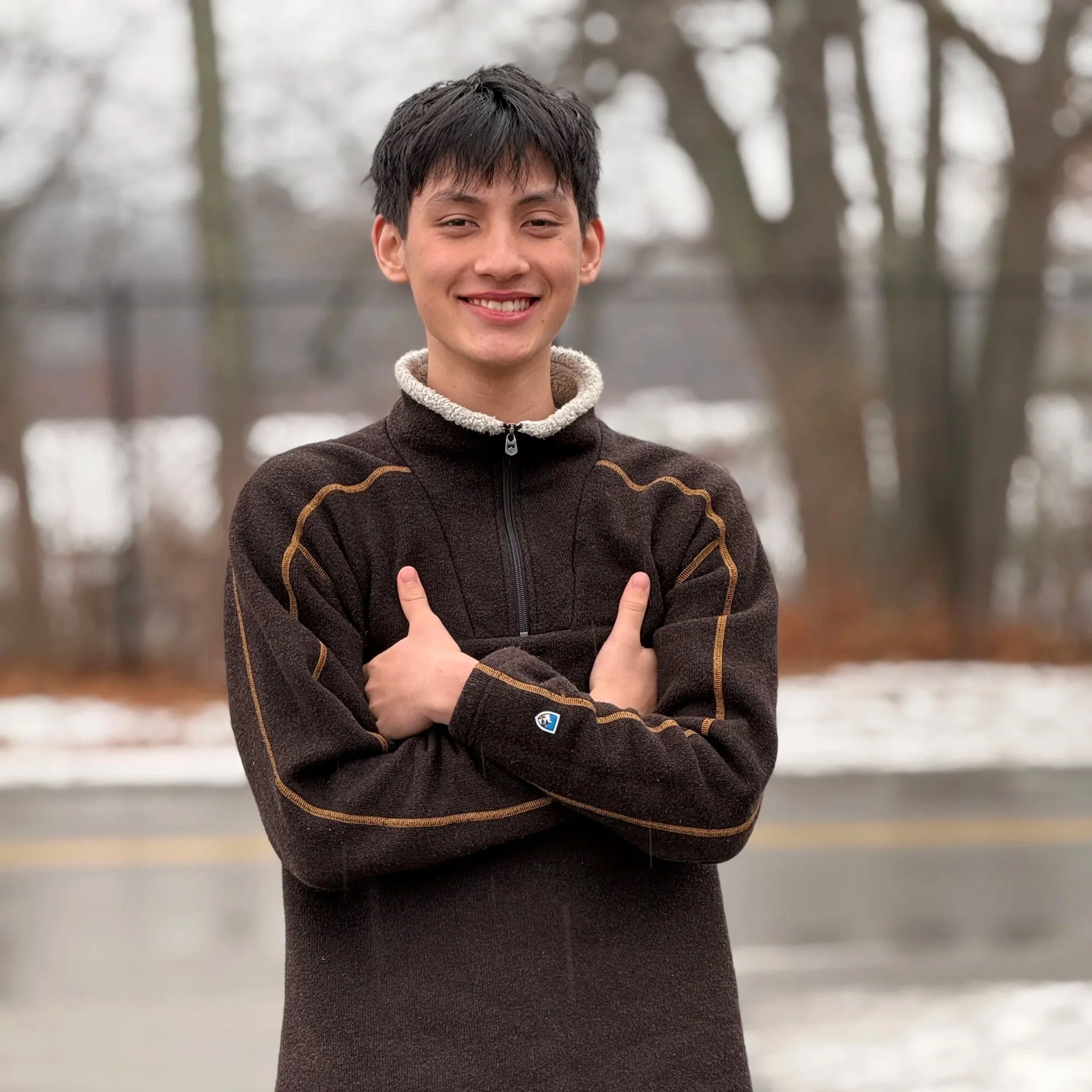 A young man with dark hair smiling outdoors during winter, wearing a dark fleece jacket with gold piping and a beige fleece collar, standing with arms crossed and crossing his arms in front of his chest.