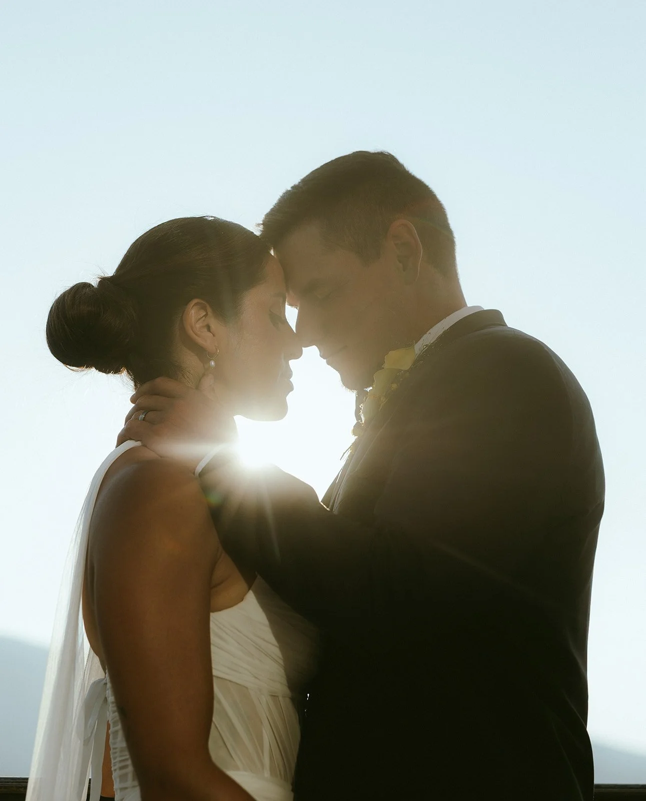 A couple in wedding attire touching foreheads and holding each other against a bright sky, backlit with sunlight.
