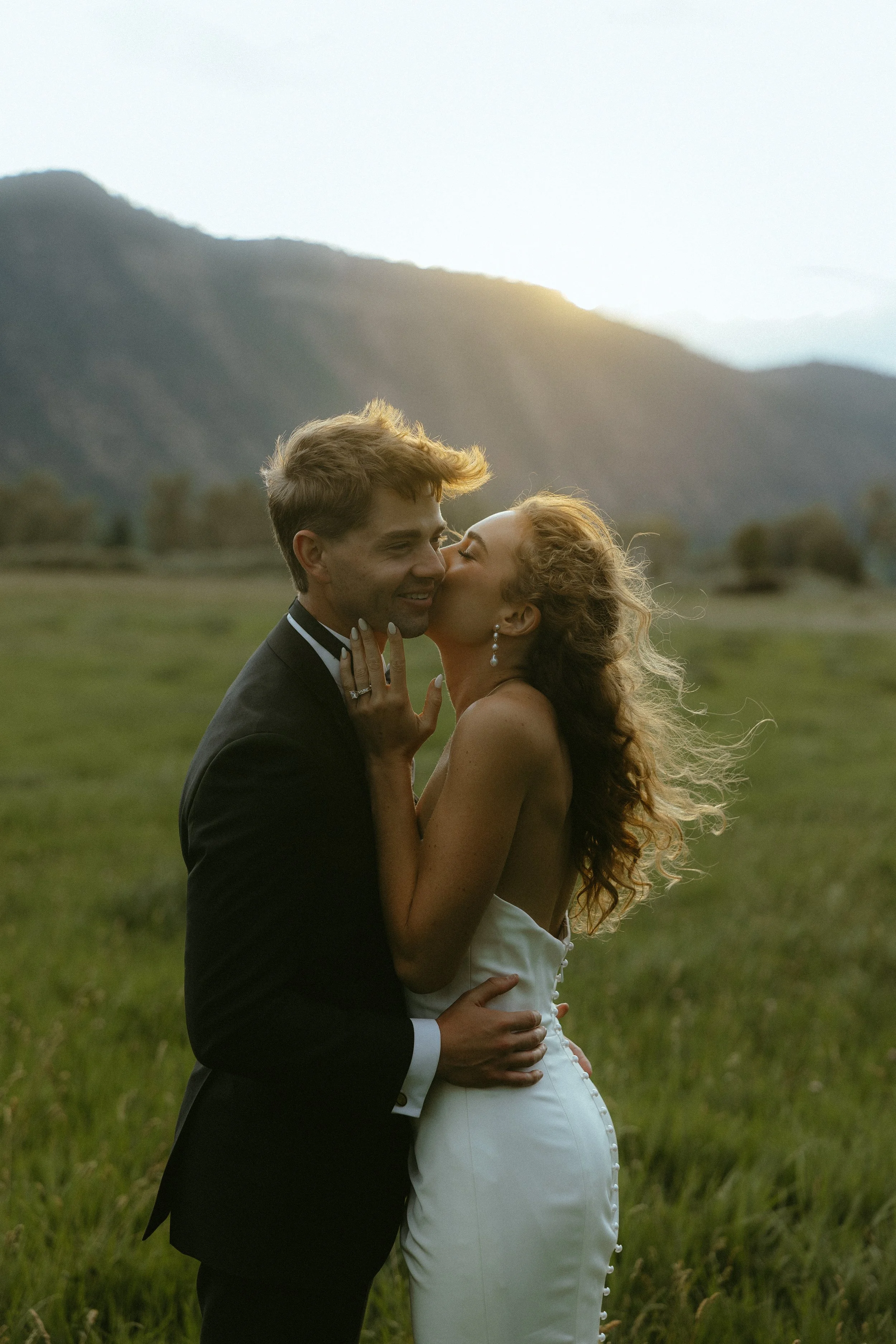 A newlywed couple sharing a kiss outdoors in a grassy field with mountains in the background during sunset.