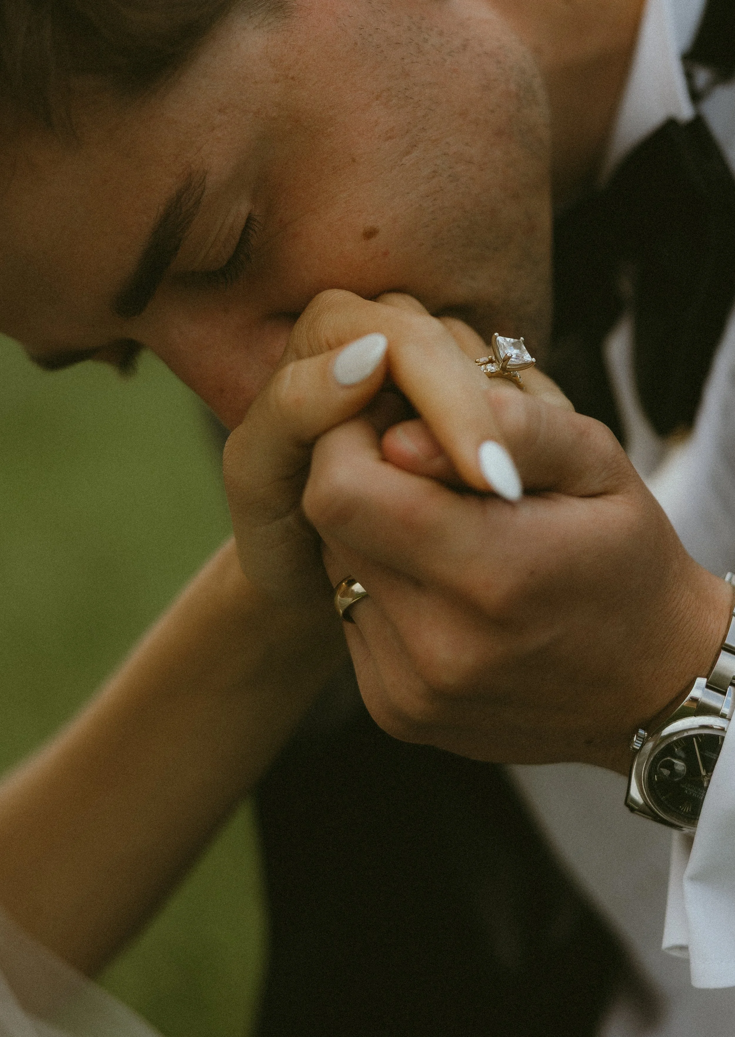 Close-up of a groom holding & kissing his wife's hands while she wears wearing a large sparkling ring, a silver watch, and white nail polish.
