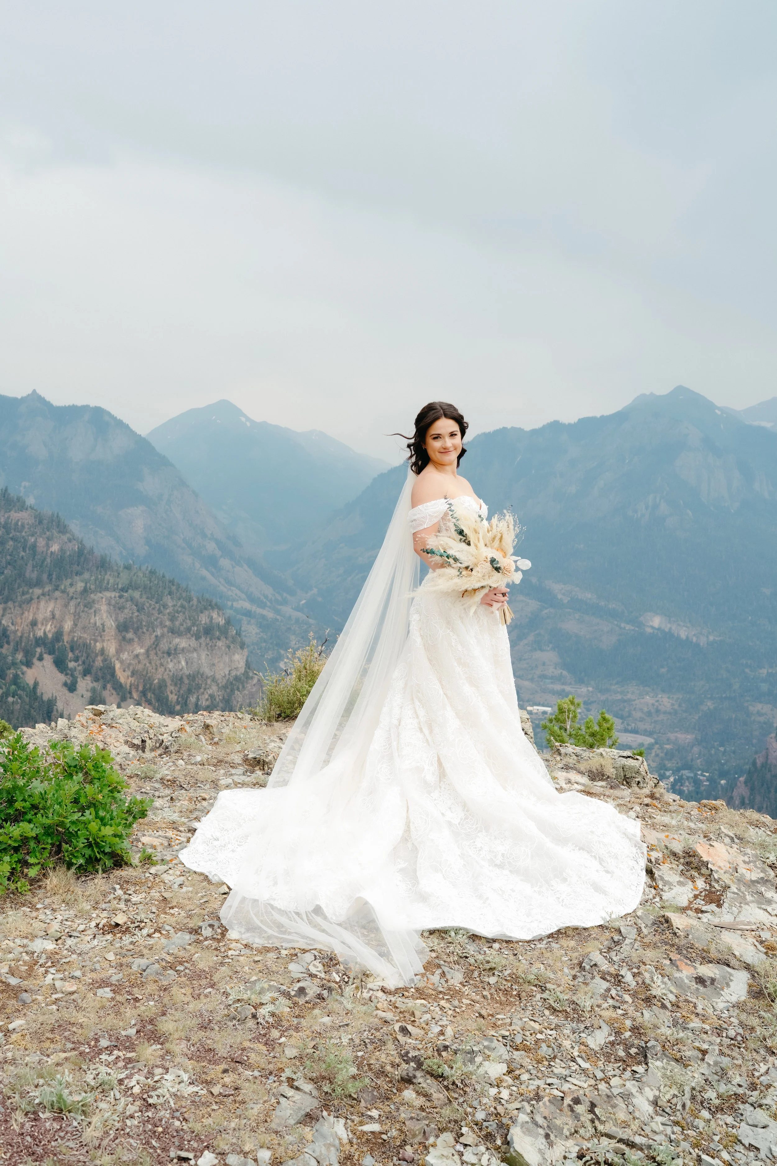 A woman in a white wedding gown holding a bouquet of flowers, standing outdoors on rocky terrain with mountain ranges in the background under a cloudy sky.