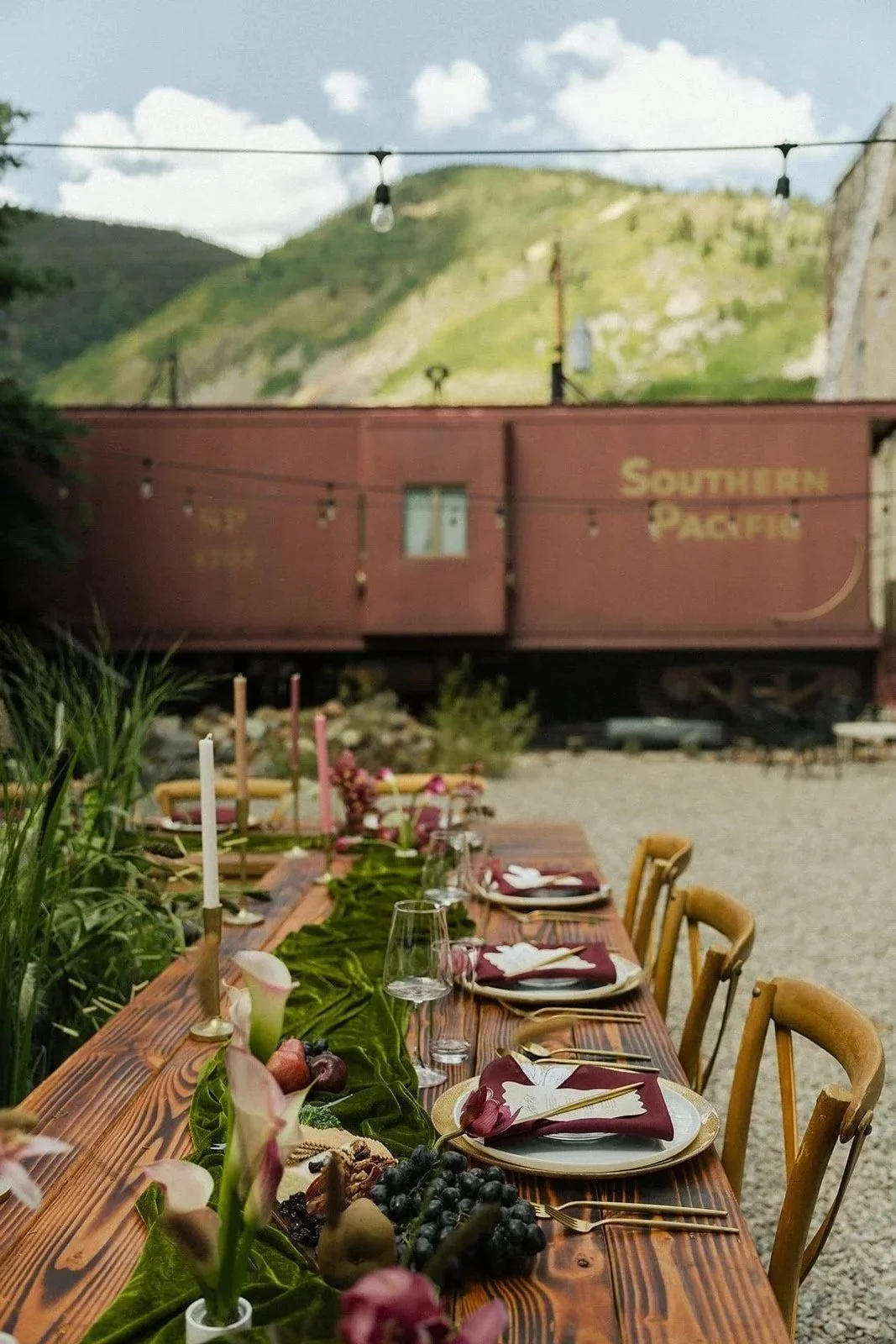 Outdoor dining table with elegant place settings, floral decorations, and candles, set against a backdrop of a hill and a vintage Southern Pacific train car.