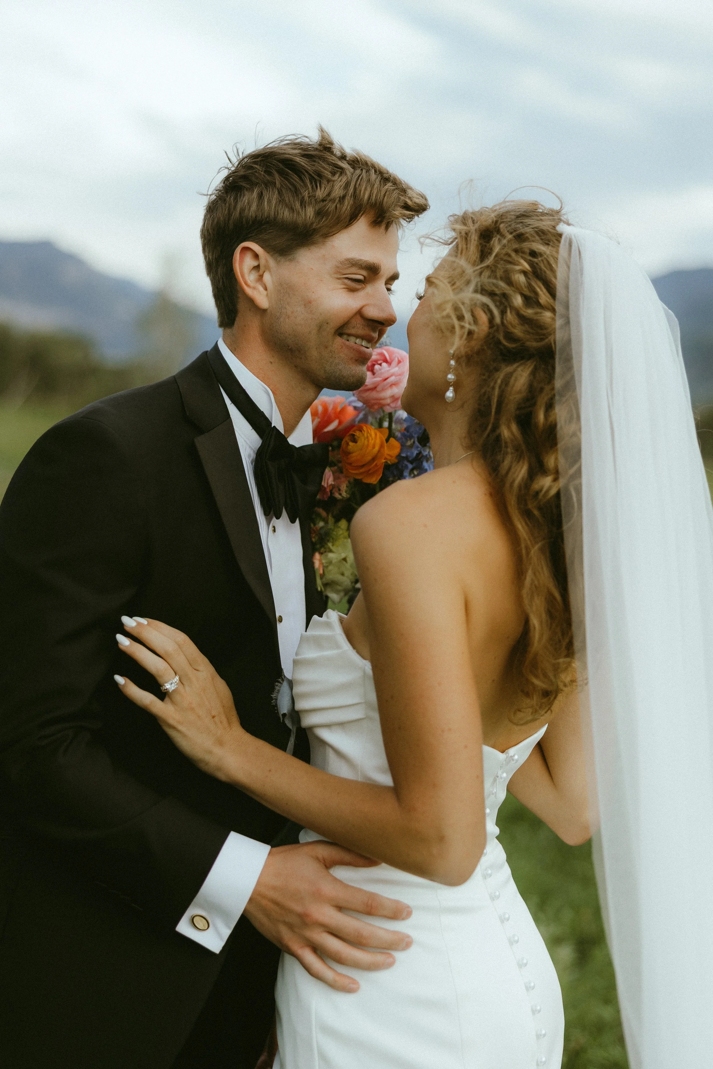 A groom and bride smiling and leaning close during their wedding outdoors, with mountains in the background.