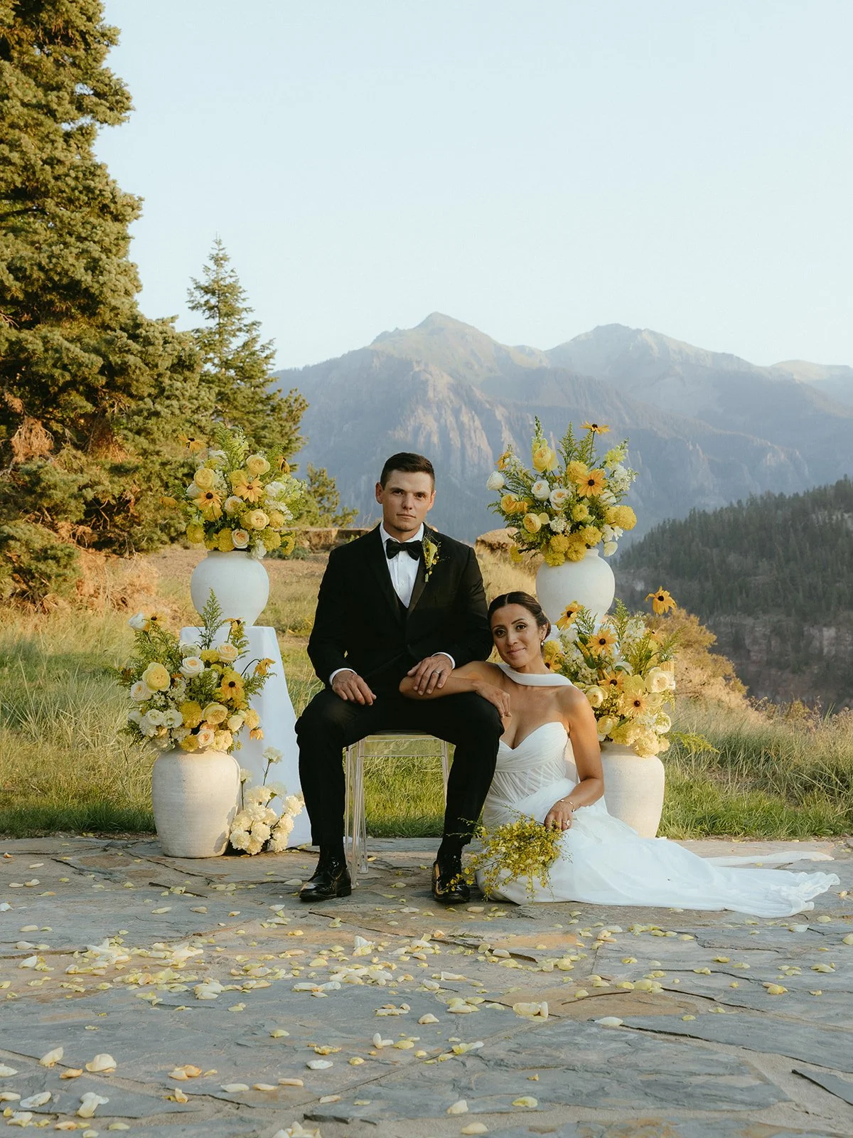 A bride and groom sitting outdoors with mountain scenery in the background. The groom is dressed in a tuxedo with a black bow tie, and the bride in a white wedding gown holding a bouquet. They are surrounded by large vases filled with yellow and white flowers, and the ground is covered with scattered flower petals.