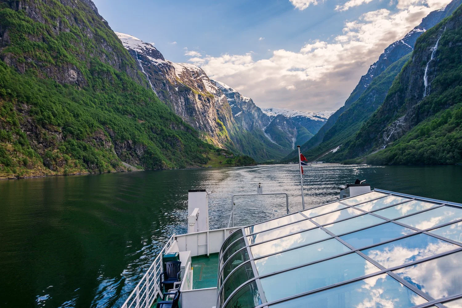 View from the bow of a cruise ship traveling through a fjord with steep, green mountains on either side and snow-capped peaks in the distance. The sky is partly cloudy with sunlight illuminating the landscape.