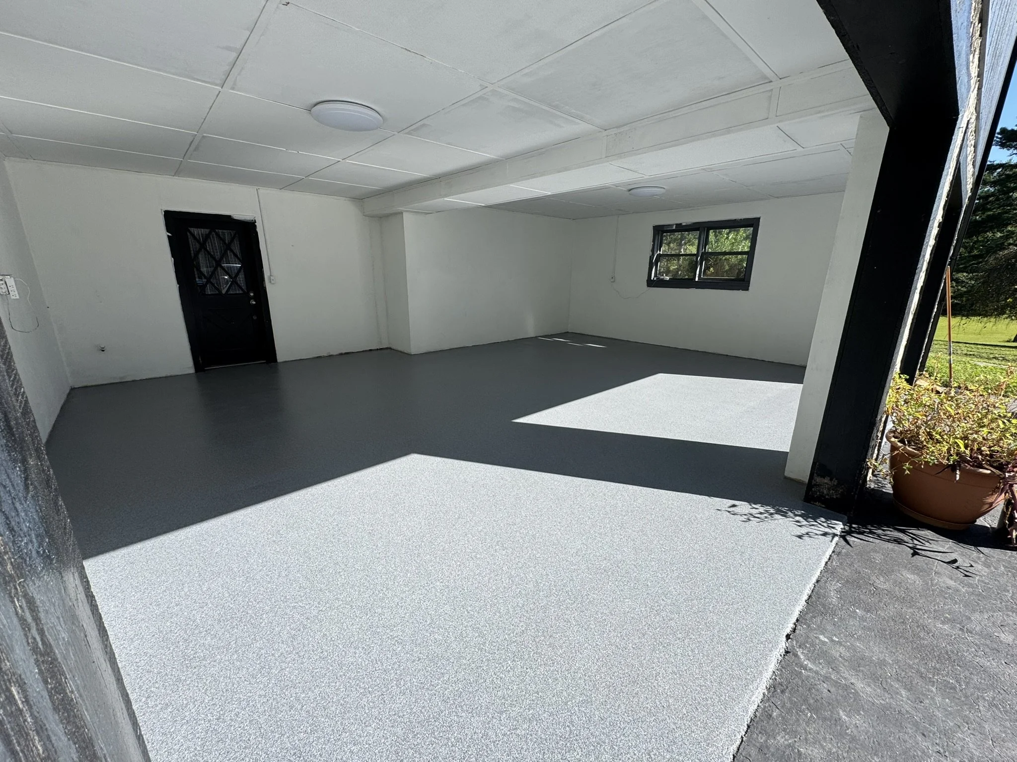 Empty indoor space with white walls, gray flooring, black door, and black window, illuminated by natural light.