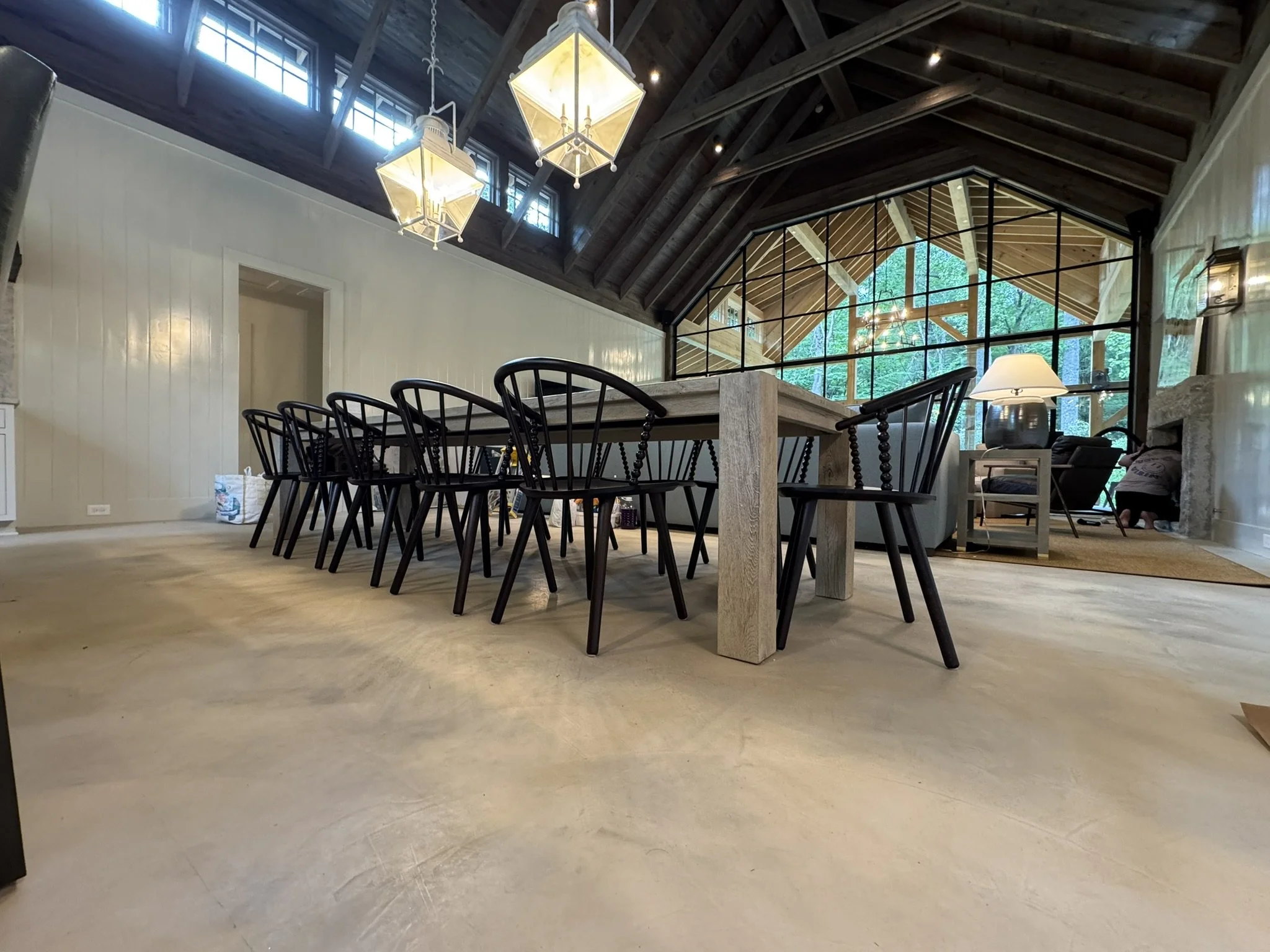 Dining room with a long wooden table and black chairs, high vaulted ceiling with hanging light fixtures, large windows and glass wall showing an outdoor wooden structure and greenery.