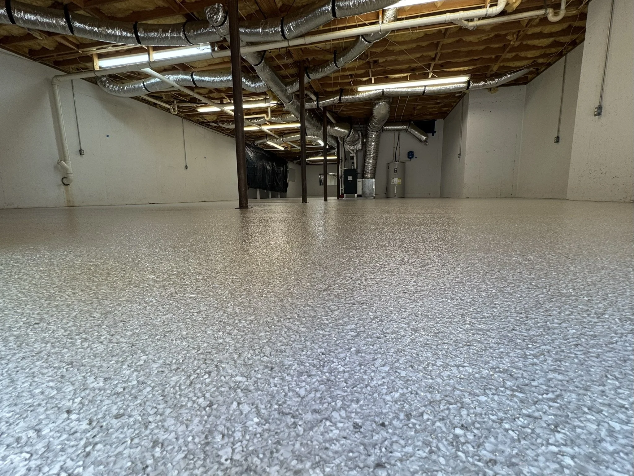 Empty basement with textured epoxy floor, exposed pipes and ductwork, and support poles.