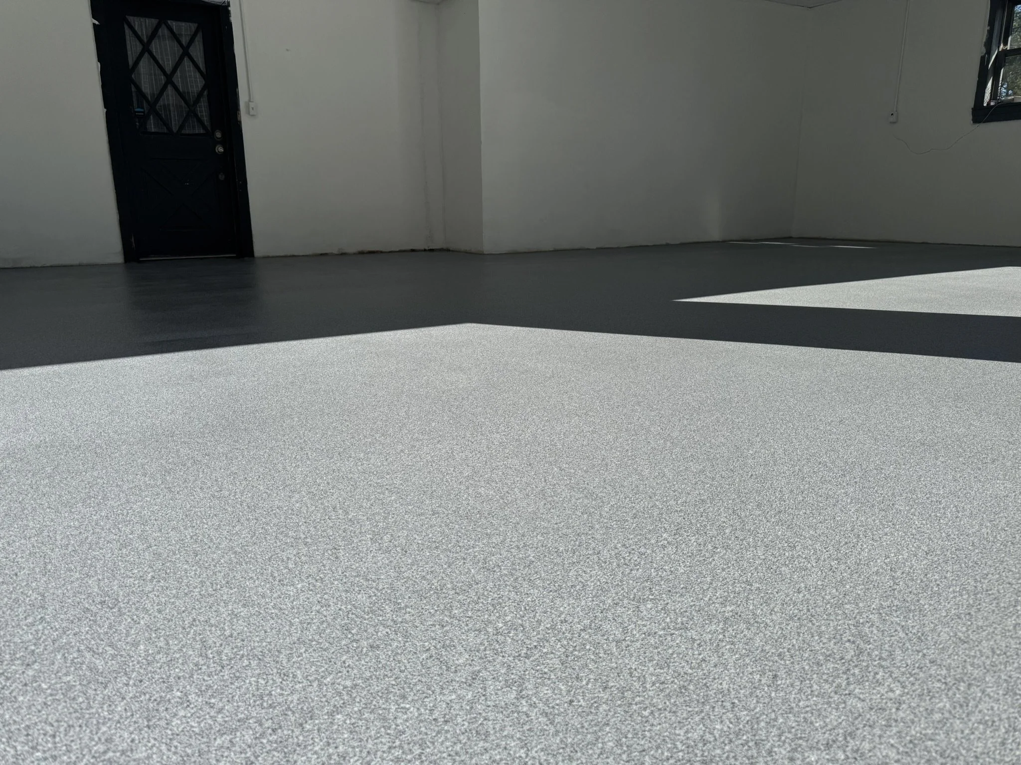 Empty room with gray flooring, white walls, black door, and windows letting in sunlight.