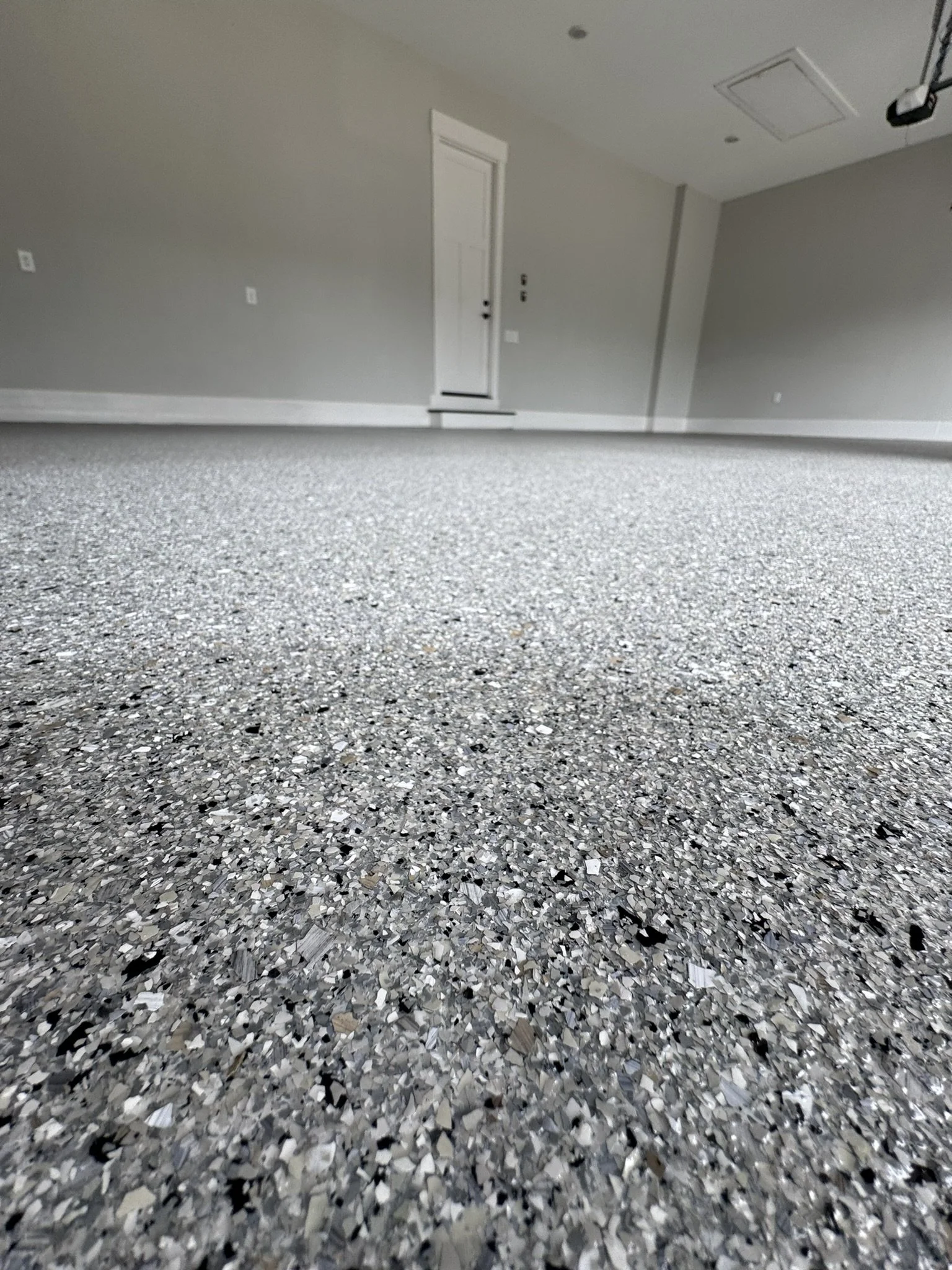 Low-angle view of a speckled granite floor in an empty room with a white door, gray walls, and ceiling with vents and a device hanging from the ceiling.
