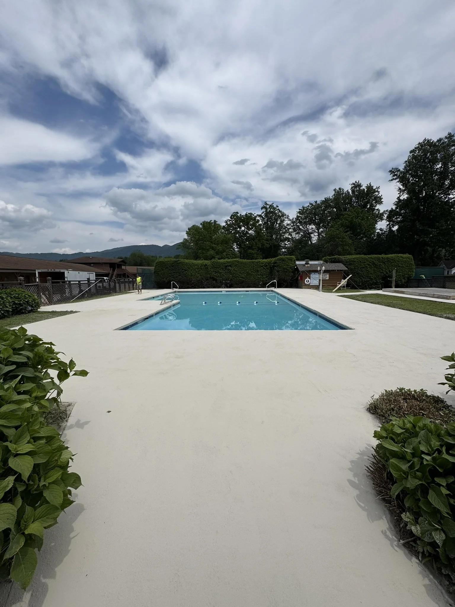 An outdoor swimming pool with a concrete pool deck, surrounded by green bushes and trees, under a partly cloudy sky with rolling hills in the background.