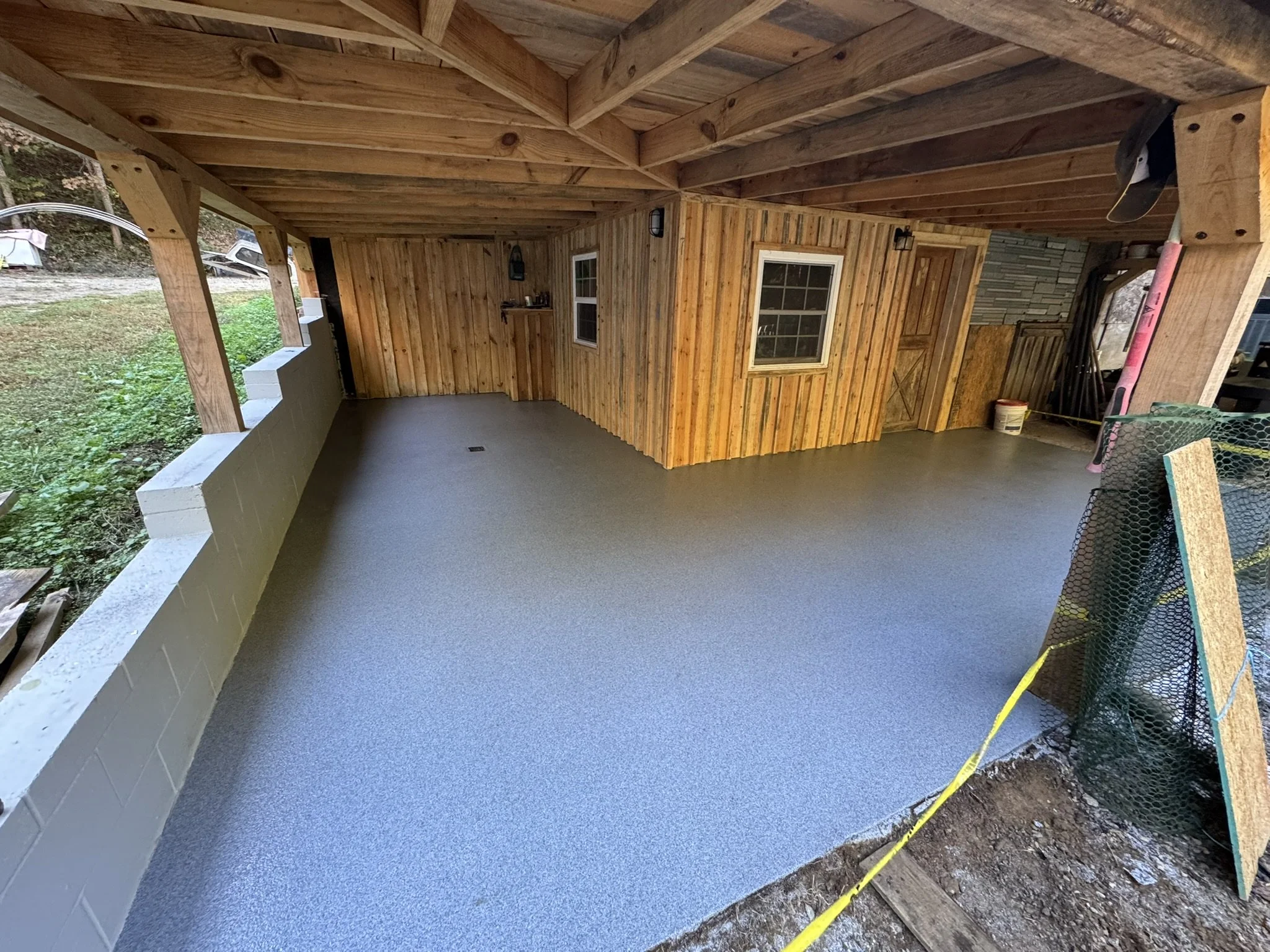 A covered porch with wood paneling and a concrete floor. There are two windows on the wood wall, and outdoor seating is partially visible on the right side. The ceiling is made of wooden beams. There is construction equipment and materials on the rig
