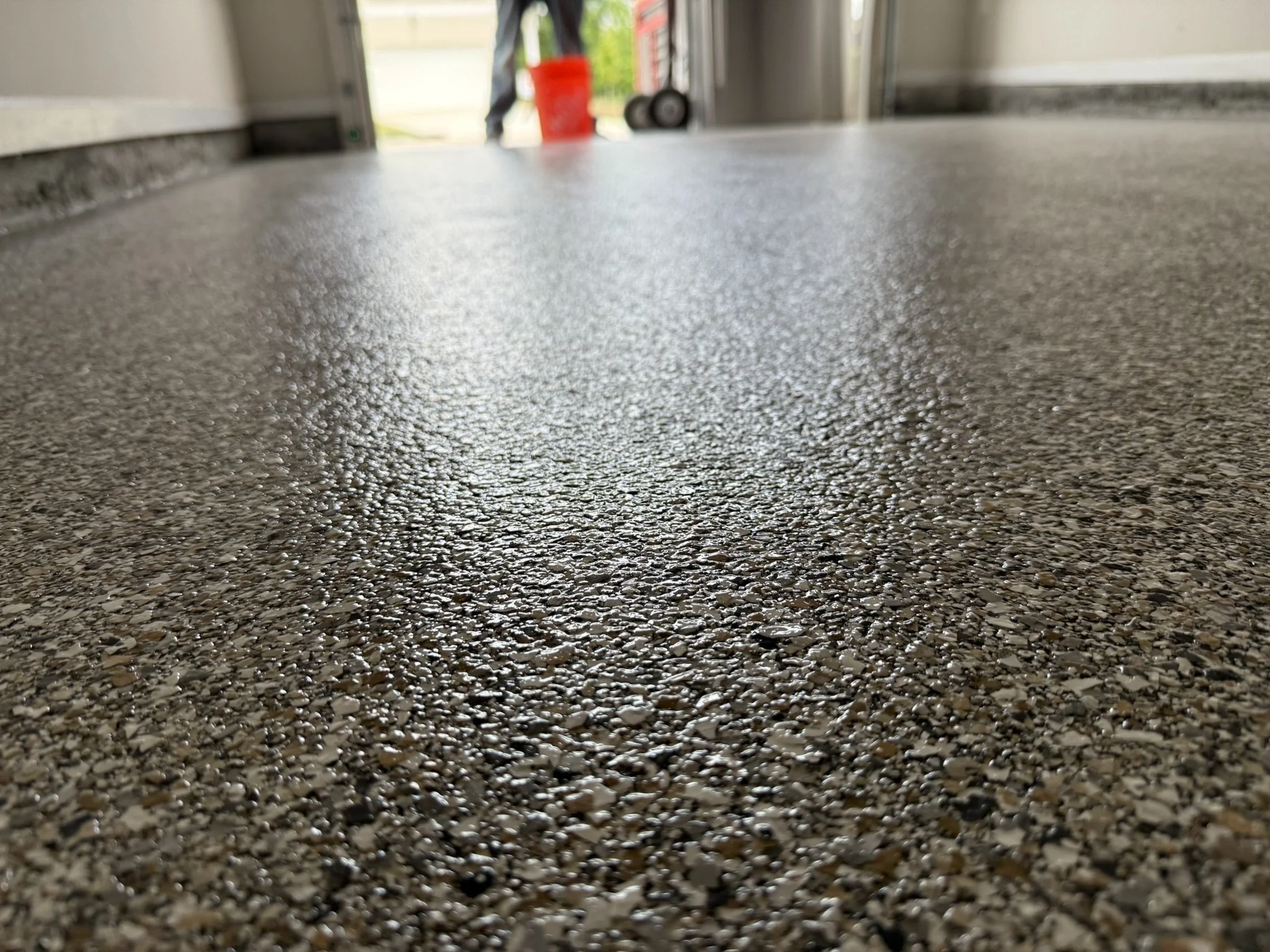 Close-up of a speckled epoxy garage floor with a slightly wet or shiny surface, showing the garage opening in the background with a red bucket and a person walking outside.