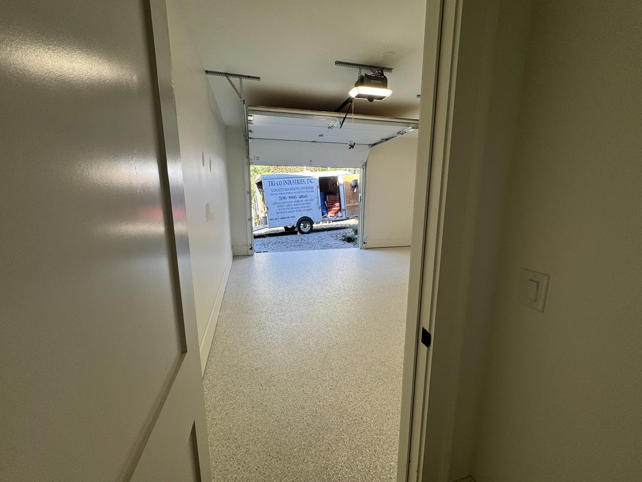 View of an open garage with a concrete floor, white walls, and a ceiling-mounted garage door opener. Outside, there is a moving truck or van parked on a gravel area with some greenery and a building in the background.