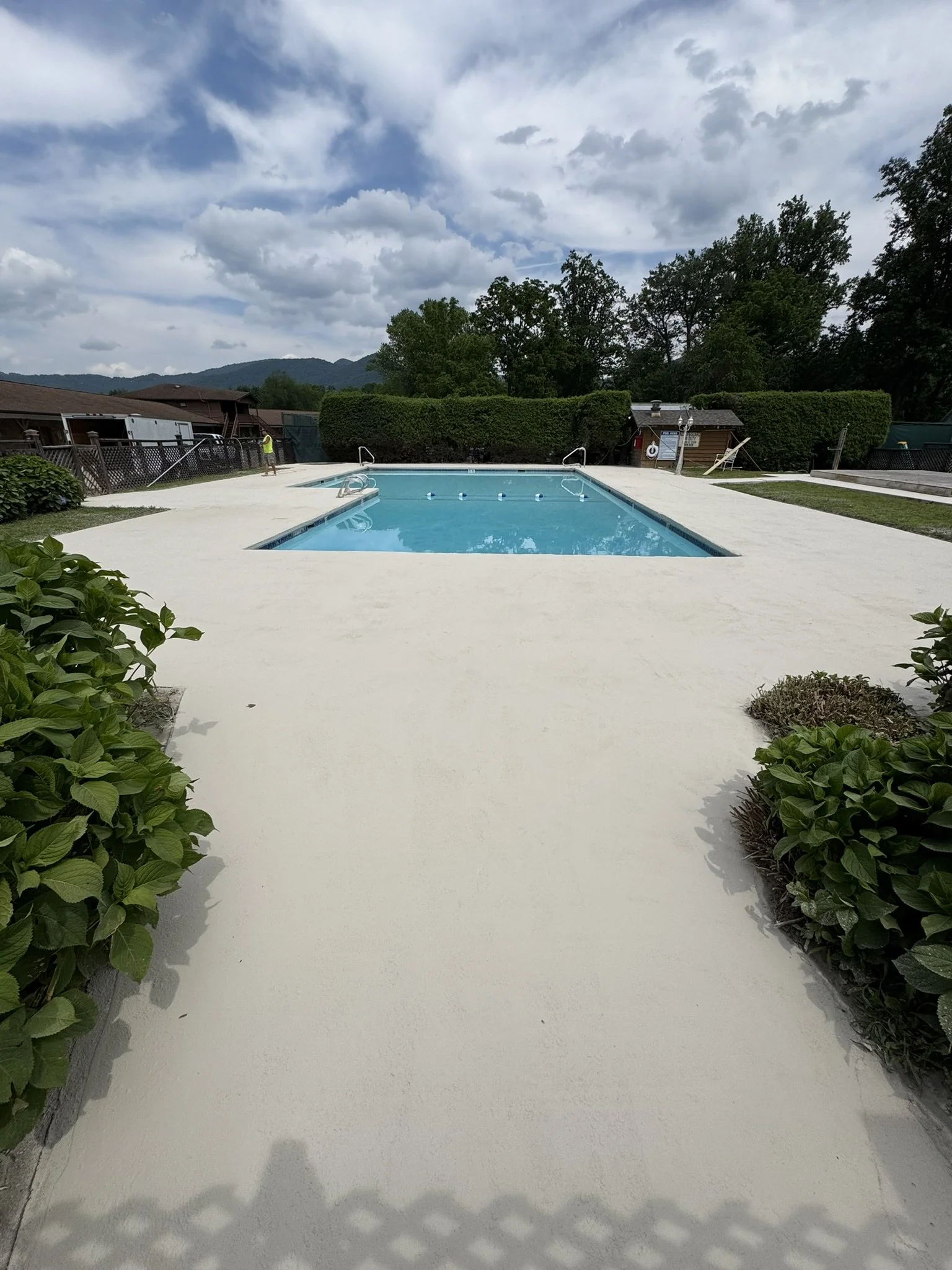 An empty swimming pool with a concrete deck, surrounded by green shrubbery and trees, under a cloudy sky with mountains in the background.