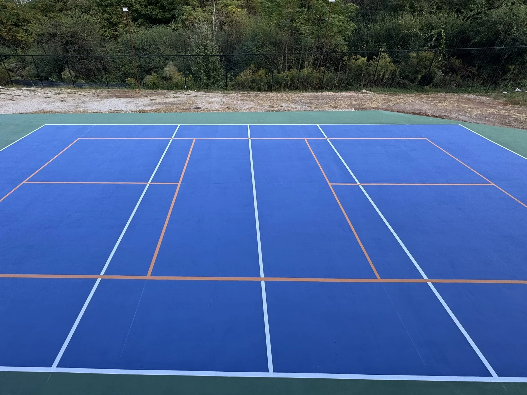An outdoor tennis or pickleball court with a blue playing surface and white lines, surrounded by trees and fencing.