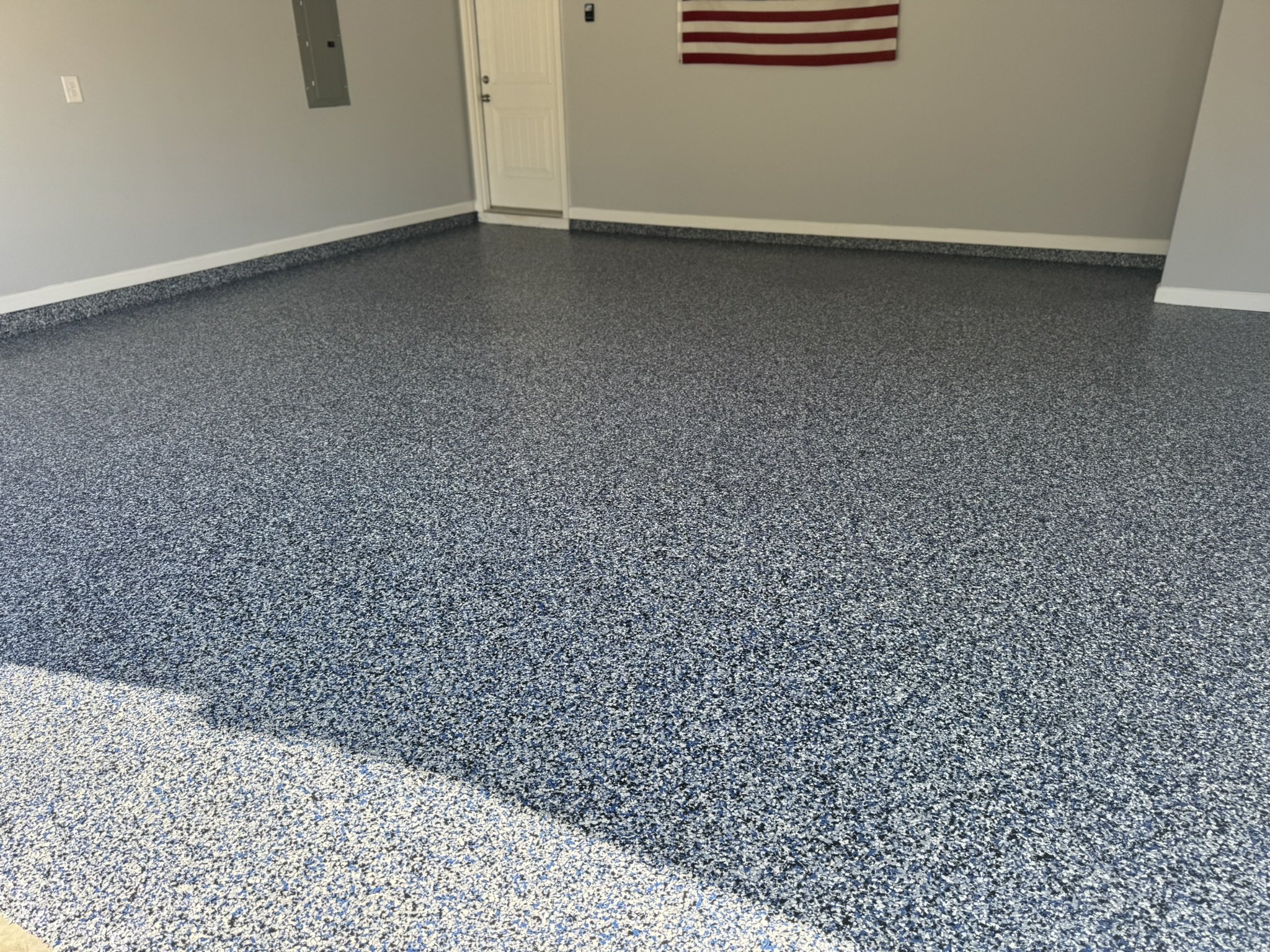 Empty garage with speckled blue, gray, and white epoxy-coated floor, neutral gray walls, American flag hanging on the wall, and a closed utility door.