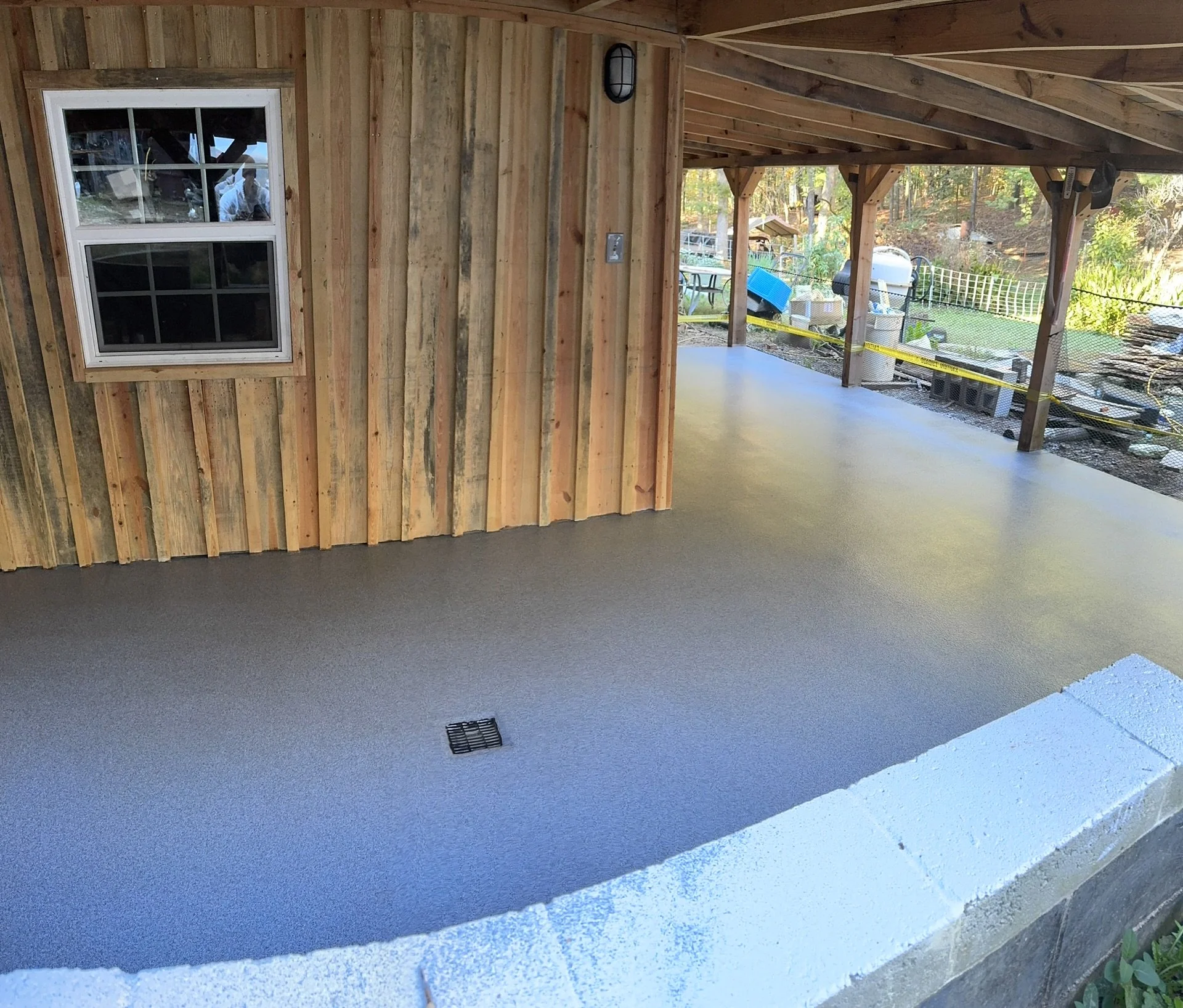 An outdoor porch area with a freshly sealed concrete floor, wood-paneled walls, a window, and supporting wooden beams. The yard beyond has trees and outdoor furniture.