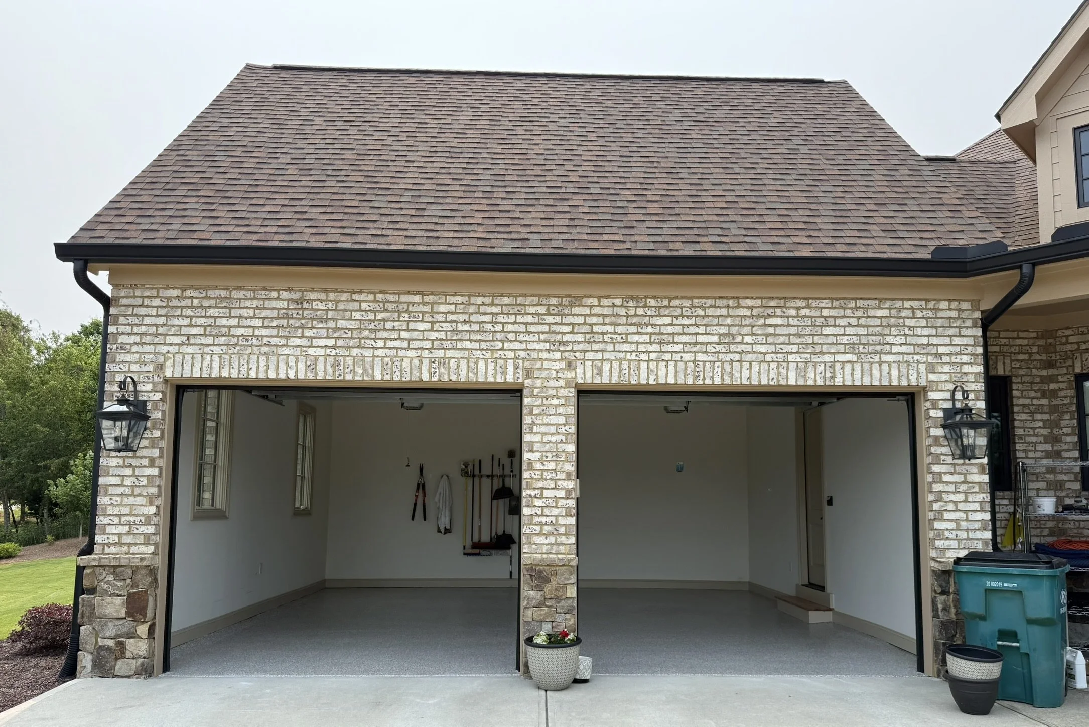 Empty two-car garage with open doors, brick exterior, and a pitched roof; tools hanging inside, potted plant outside, and a trash bin nearby.