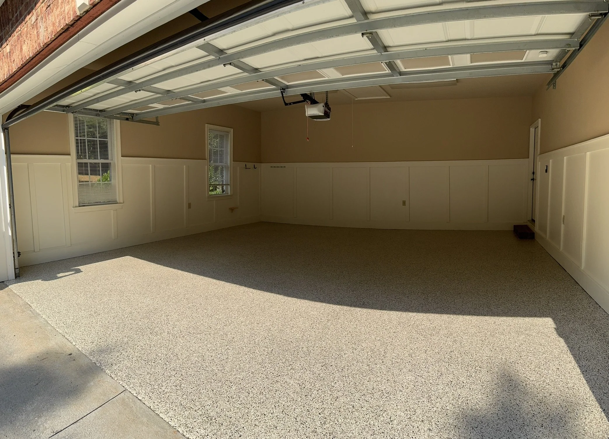 Empty residential garage with beige walls, white wainscoting, two windows, carpeted floor, and an open garage door.