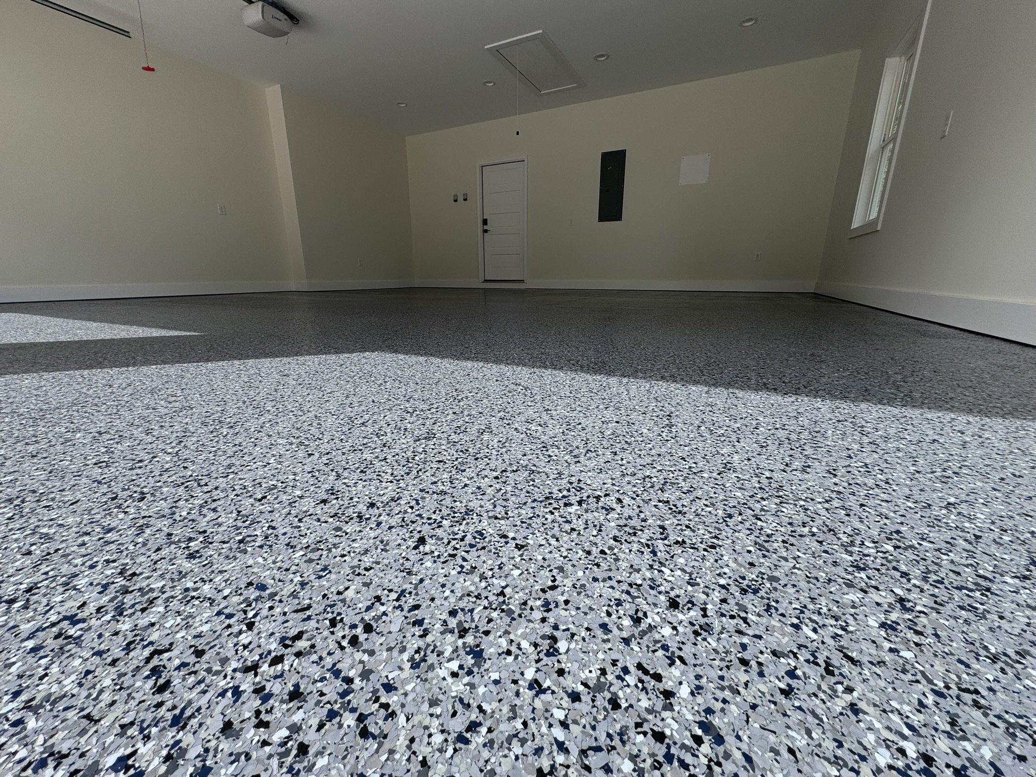 Empty room with a speckled gray, black, and white epoxy floor, cream walls, a window on the right, and a white door in the back.