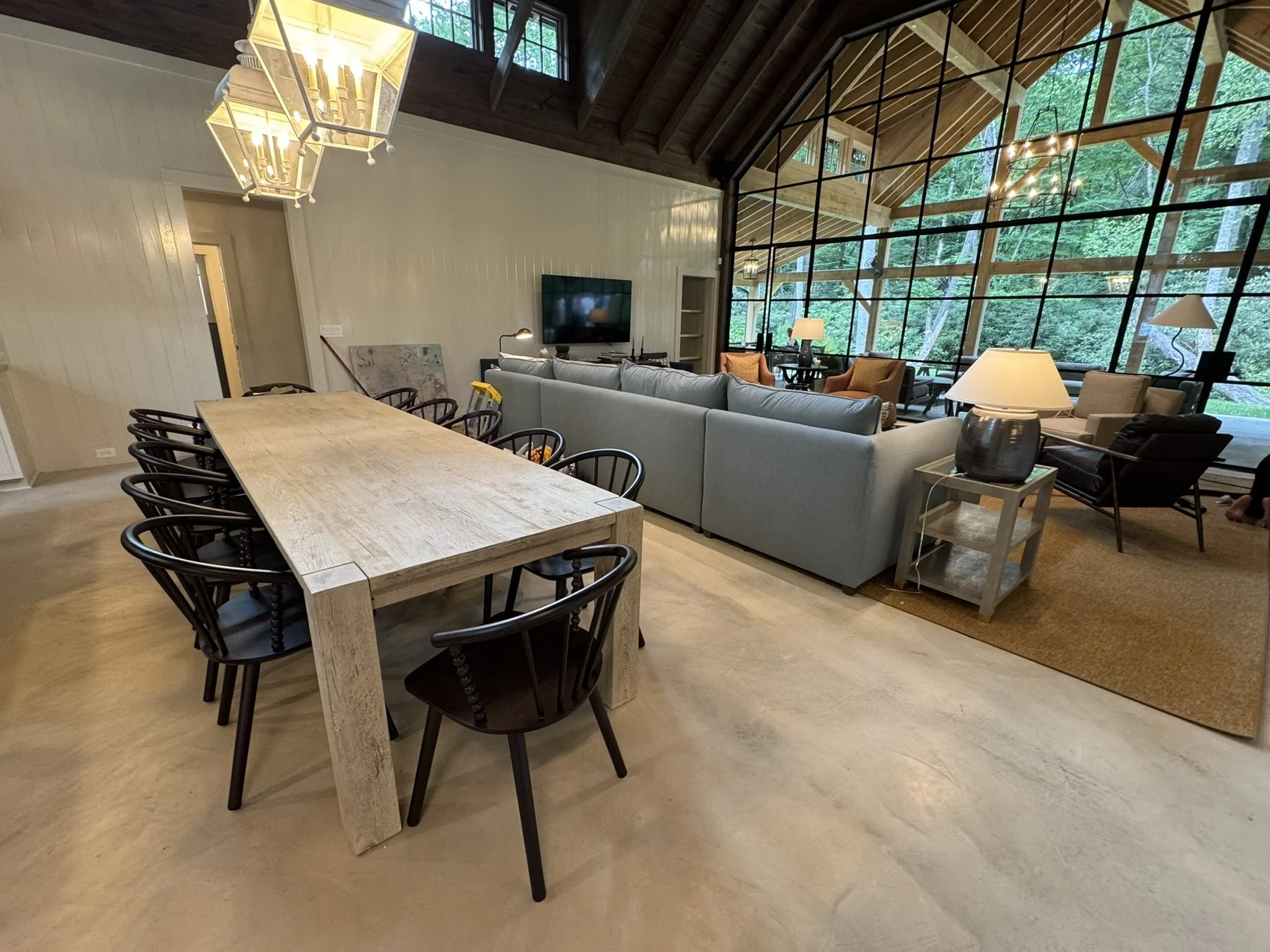 Open-concept living and dining room with a large wooden dining table surrounded by black chairs, a gray sectional sofa, armchairs, lamps, and a grid-style glass wall with views of greenery outside.