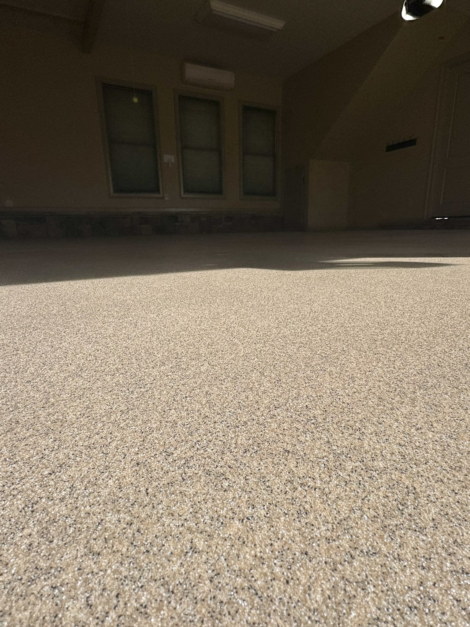 A beige outdoor patio with a textured surface, in front of a house wall with three windows, an air conditioning unit, and a door, partially in shadow.
