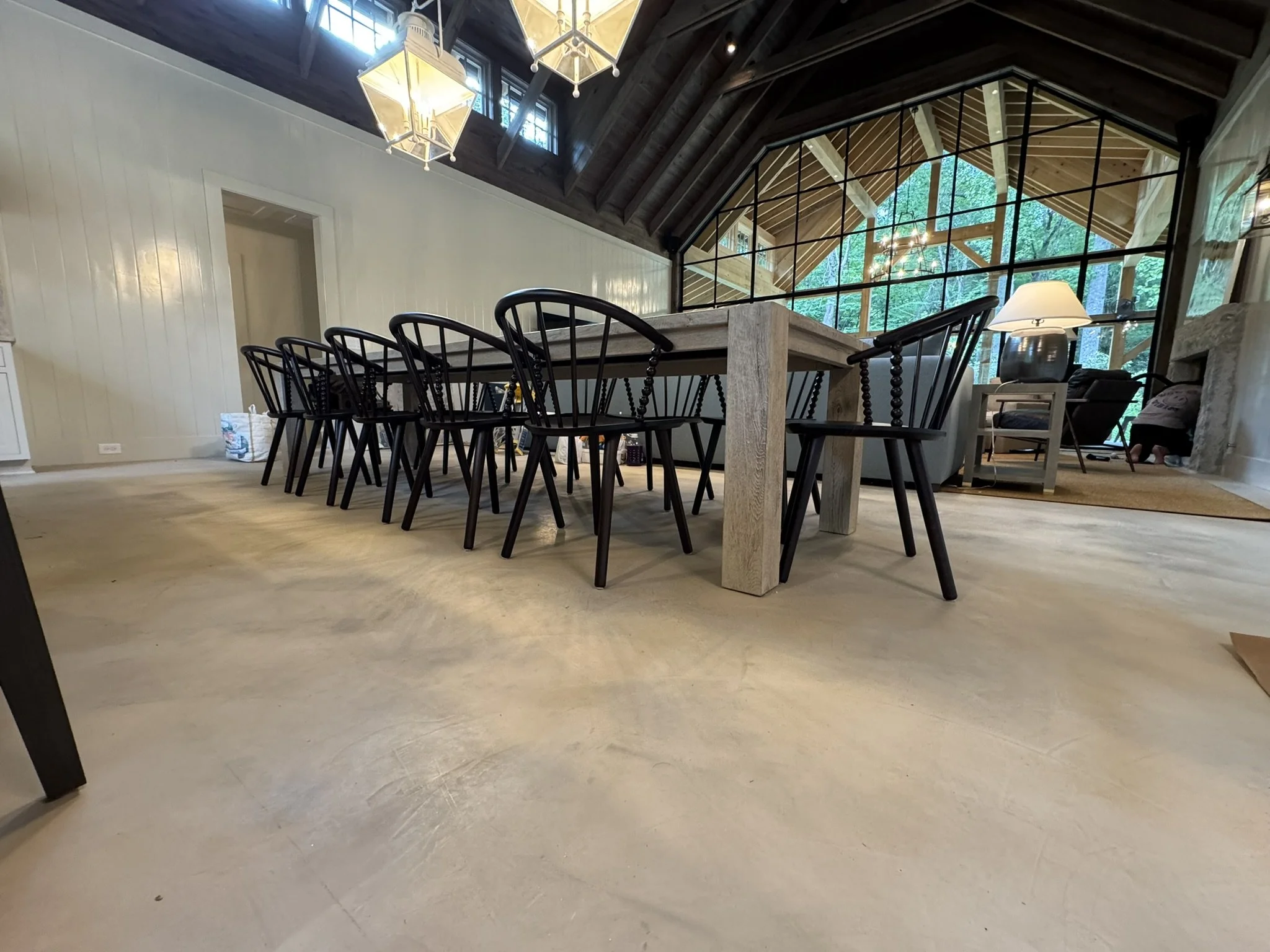 Inside view of a modern home with a dining area featuring a large rectangular wooden table, black chairs, and a living space with a lamp, a sofa, and large windows with a view of greenery outside. The ceiling has dark wood beams and a chandelier, and
