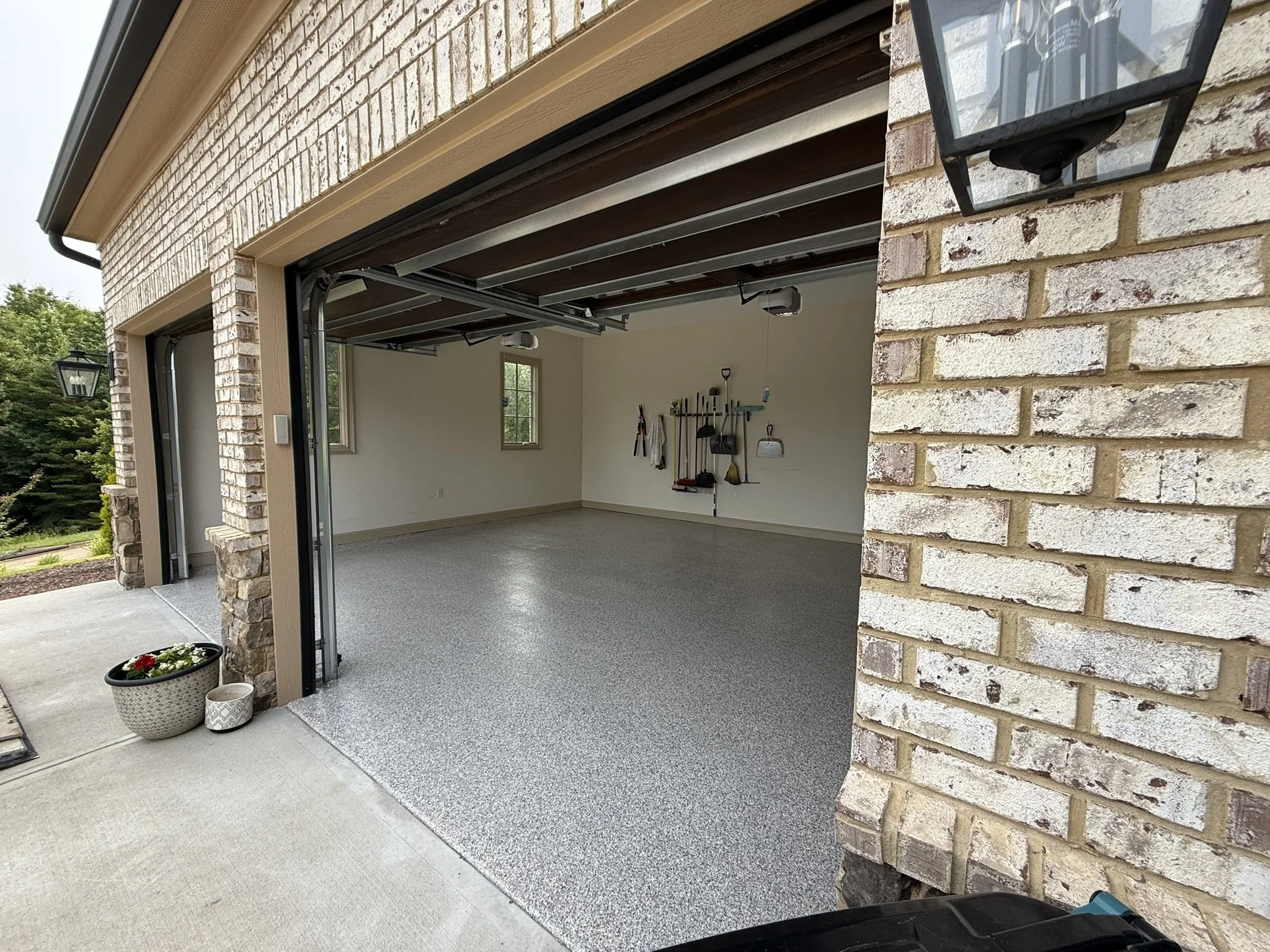 Empty residential garage with open door, pebbled floor, brick walls, window, and wall-mounted tools.