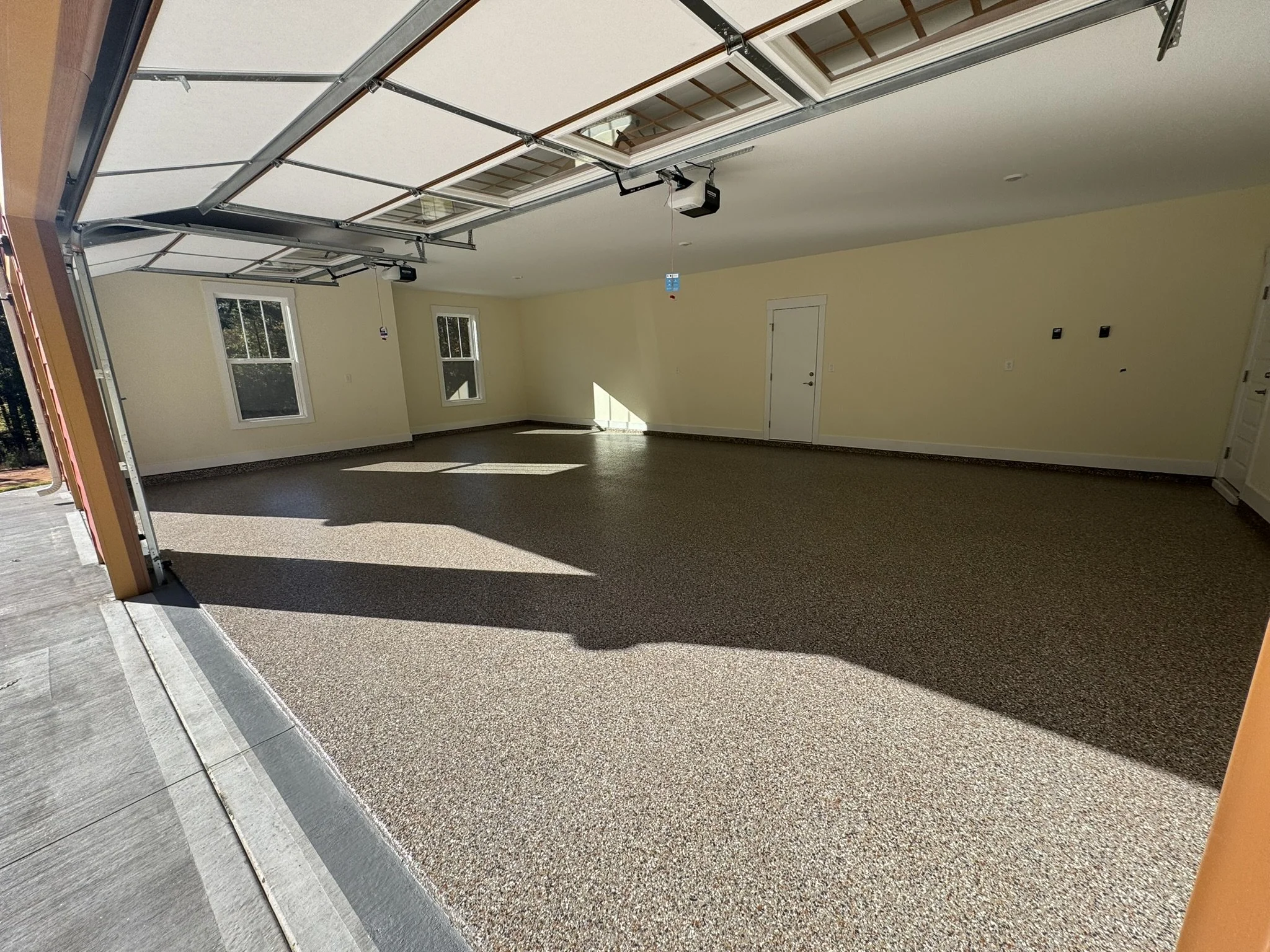 Empty residential garage with open door, concrete floor, beige walls, and two windows, sunlight casting shadows on the floor.