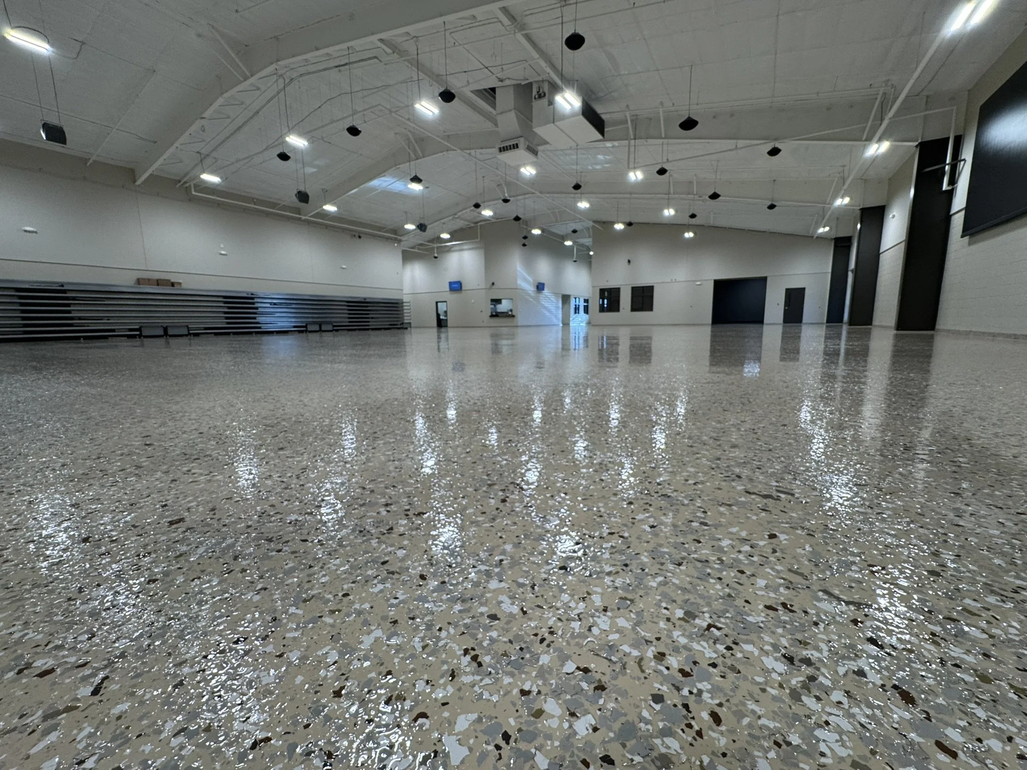 Empty indoor gymnasium with white walls, polished terrazzo floor, suspended ceiling lights, and black wall-mounted speakers.