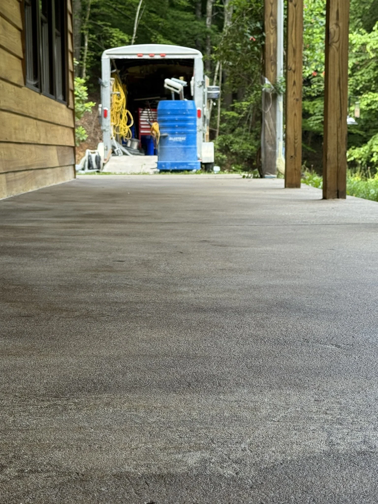 A view of a backyard porch with a textured concrete floor, a wooden wall on the left, and a wooden railing on the right. In the background, there's a trailer filled with outdoor equipment, including a yellow hose, a blue water container, and other to