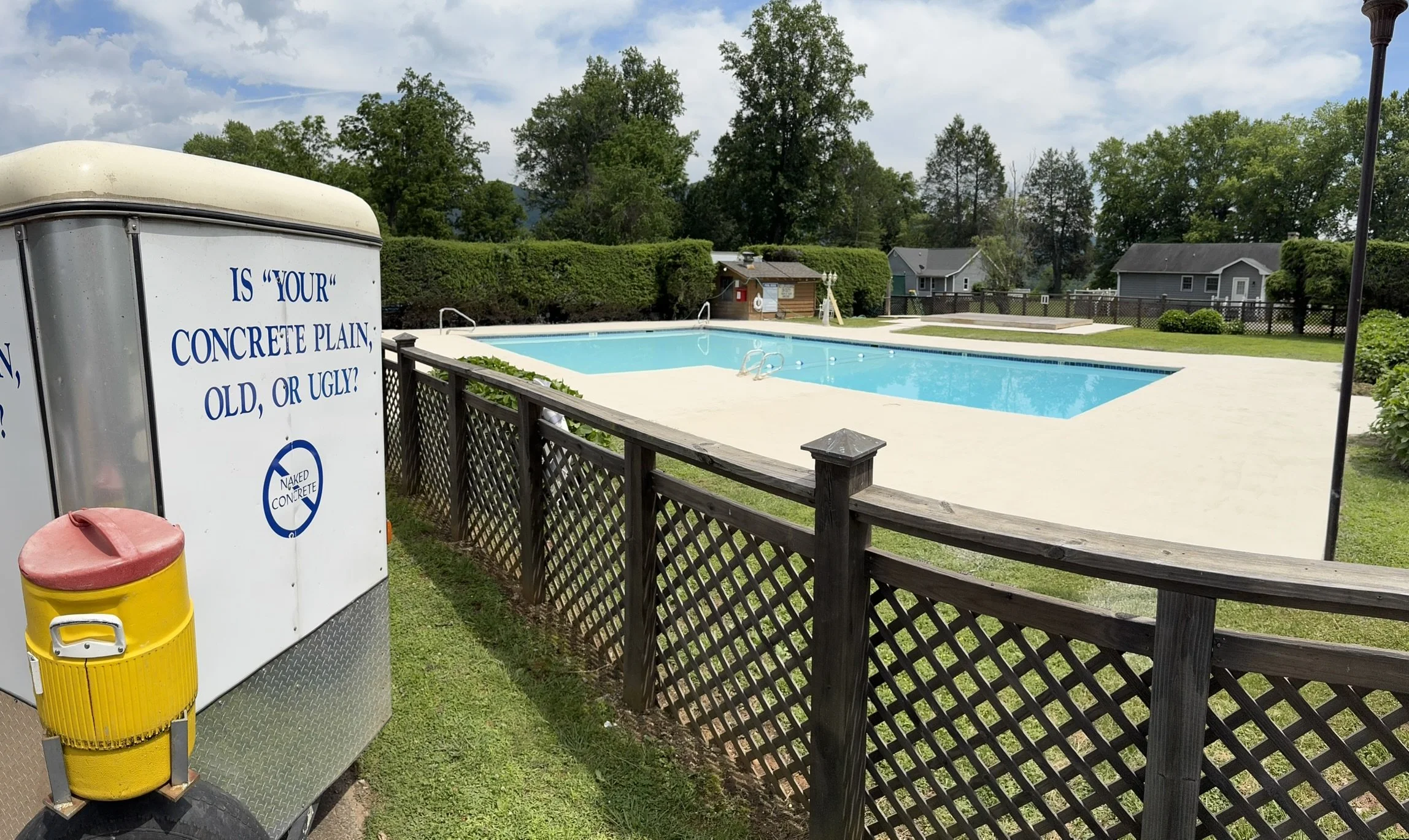 An outdoor swimming pool with a white concrete border, surrounded by a black wooden fence, with a small building and houses in the background. There is a sign on a trailer that reads, 'Is your concrete plain, old, or ugly?' and a yellow trash can nea