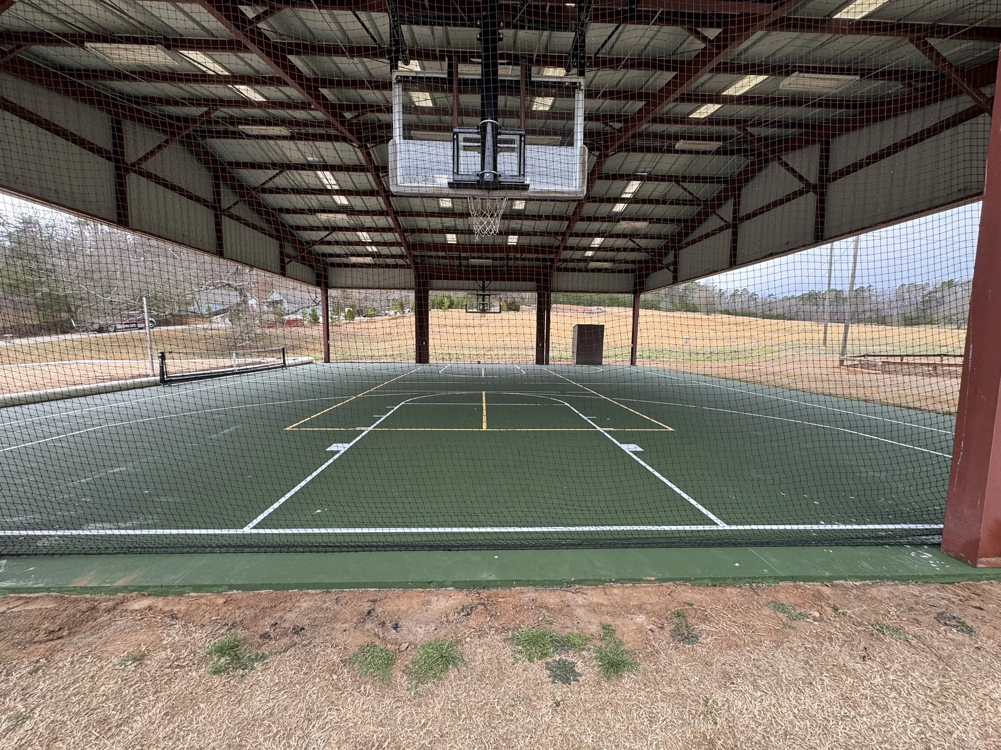 An outdoor basketball court under a large metal roof, with a basketball hoop and backboard in view. The court is surrounded by a safety net and has a dirt area outside the playing surface.