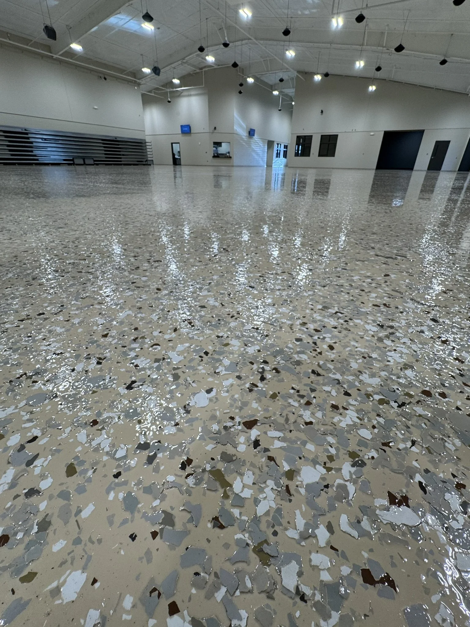 Empty indoor gymnasium with polished speckled terrazzo floor, white walls, high ceiling with hanging black acoustic panels, and a few wall-mounted TVs.