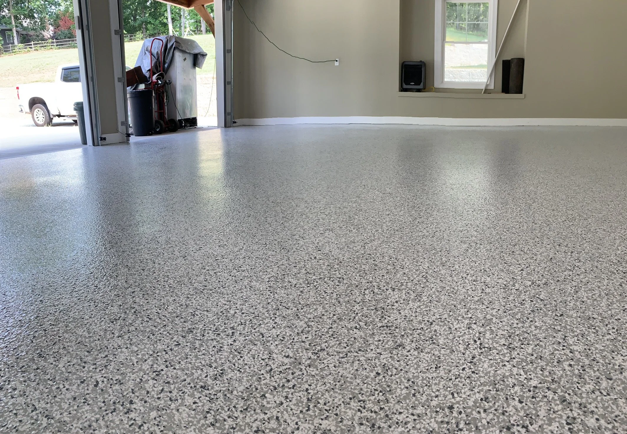 Empty garage with a textured gray floor, an open garage door showing a yard outside, and some equipment and boxes inside.
